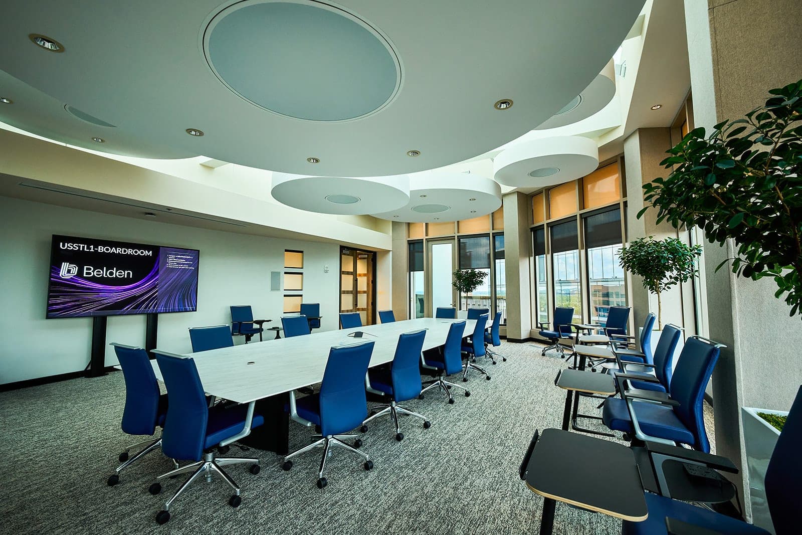 A Belden boardroom with a wall-mounted display carrying the room name and brand mark, amber window shades drawn against the afternoon light, workplace photography in St. Louis