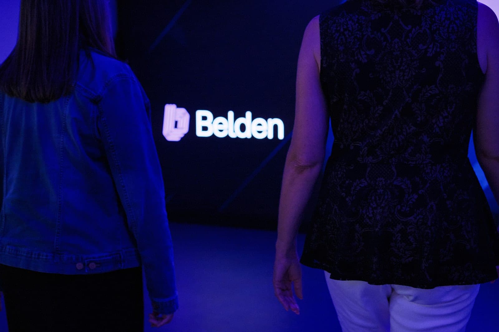 Two people walking past the illuminated Belden wordmark in a dark corridor, seen from behind, office photography in St. Louis