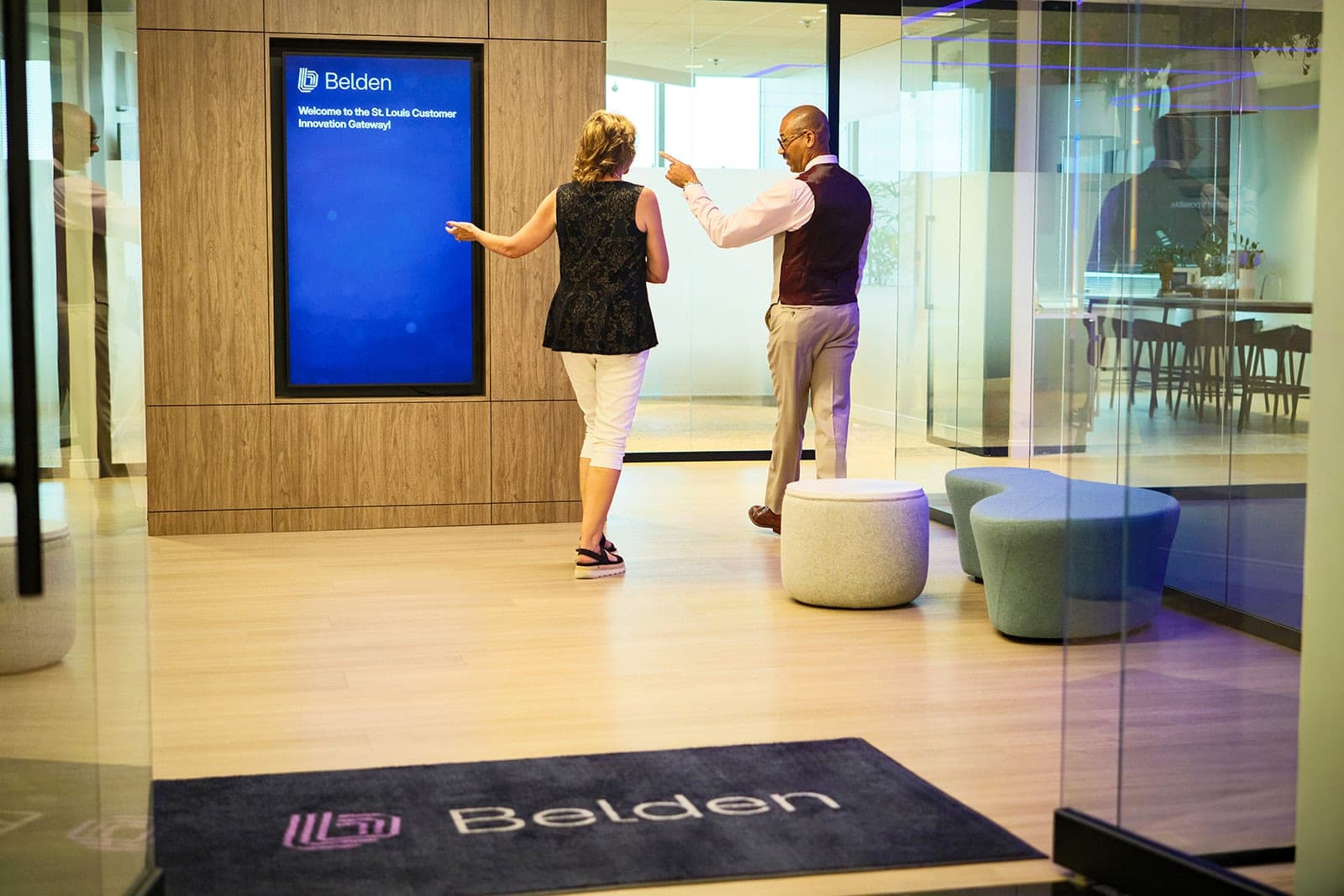 Two visitors walking away from the camera toward the welcome display at the Belden Customer Innovation Gateway, a branded floor rug in the foreground, corporate photography in St. Louis