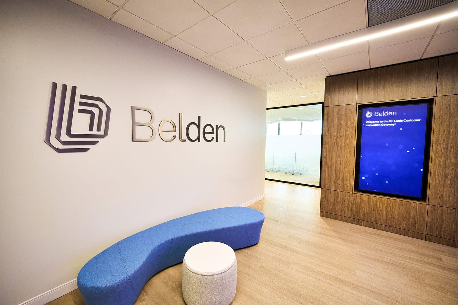 Reception at the Belden Customer Innovation Gateway in St. Louis, the dimensional Belden wordmark on a white wall beside a curved blue bench and a wood-panelled welcome display, corporate interior photography by Henry David Photography
