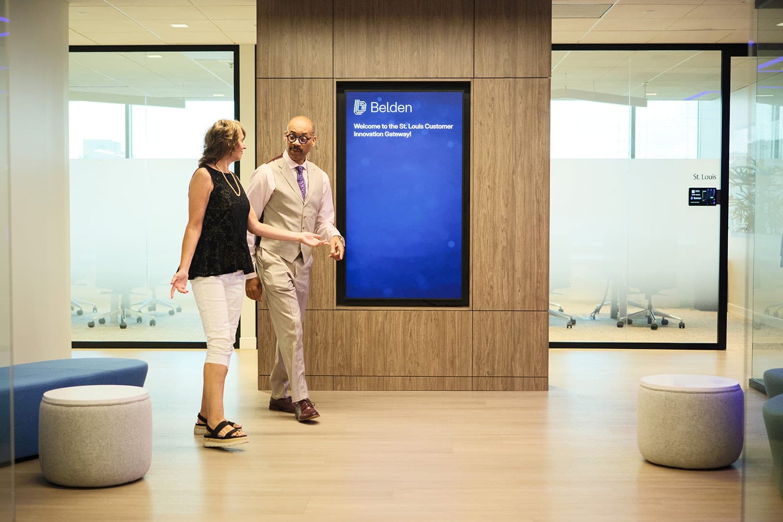 Two colleagues walking toward the illuminated welcome display at the entrance to the Belden Customer Innovation Gateway, glass-walled meeting rooms on either side, workplace photography in St. Louis