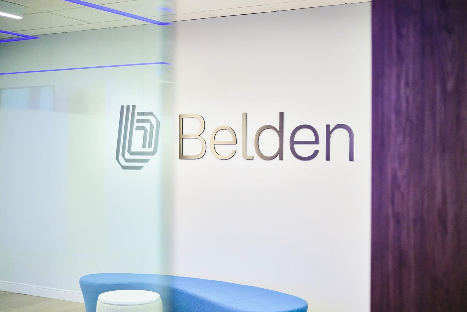 Close frame of the dimensional Belden logo and wordmark mounted on a white wall above a curved blue bench, brand detail photography in St. Louis