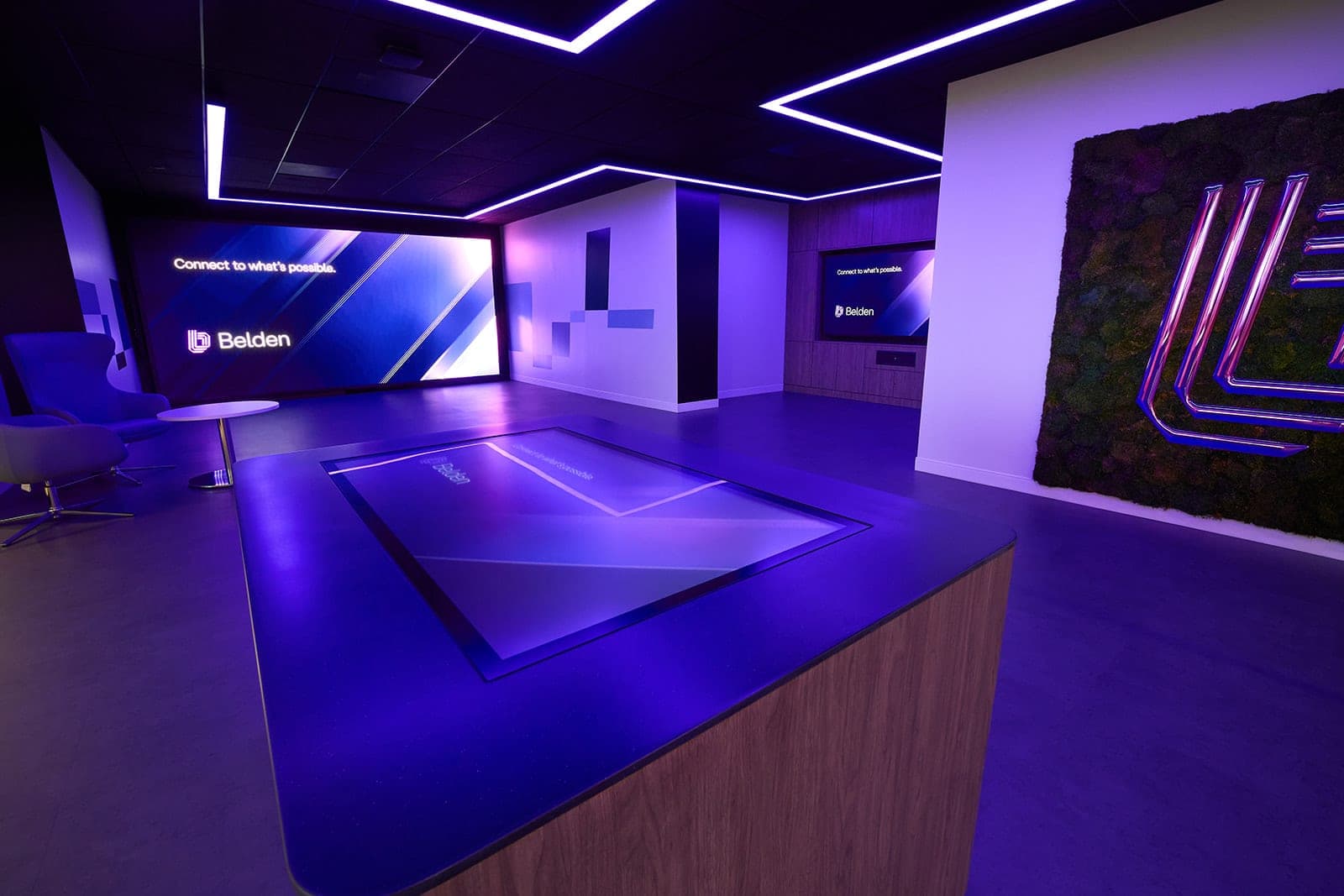 Looking past the illuminated touch table toward the moss wall and the corridor beyond it in the Belden experience center, interior photography in low ambient light in St. Louis