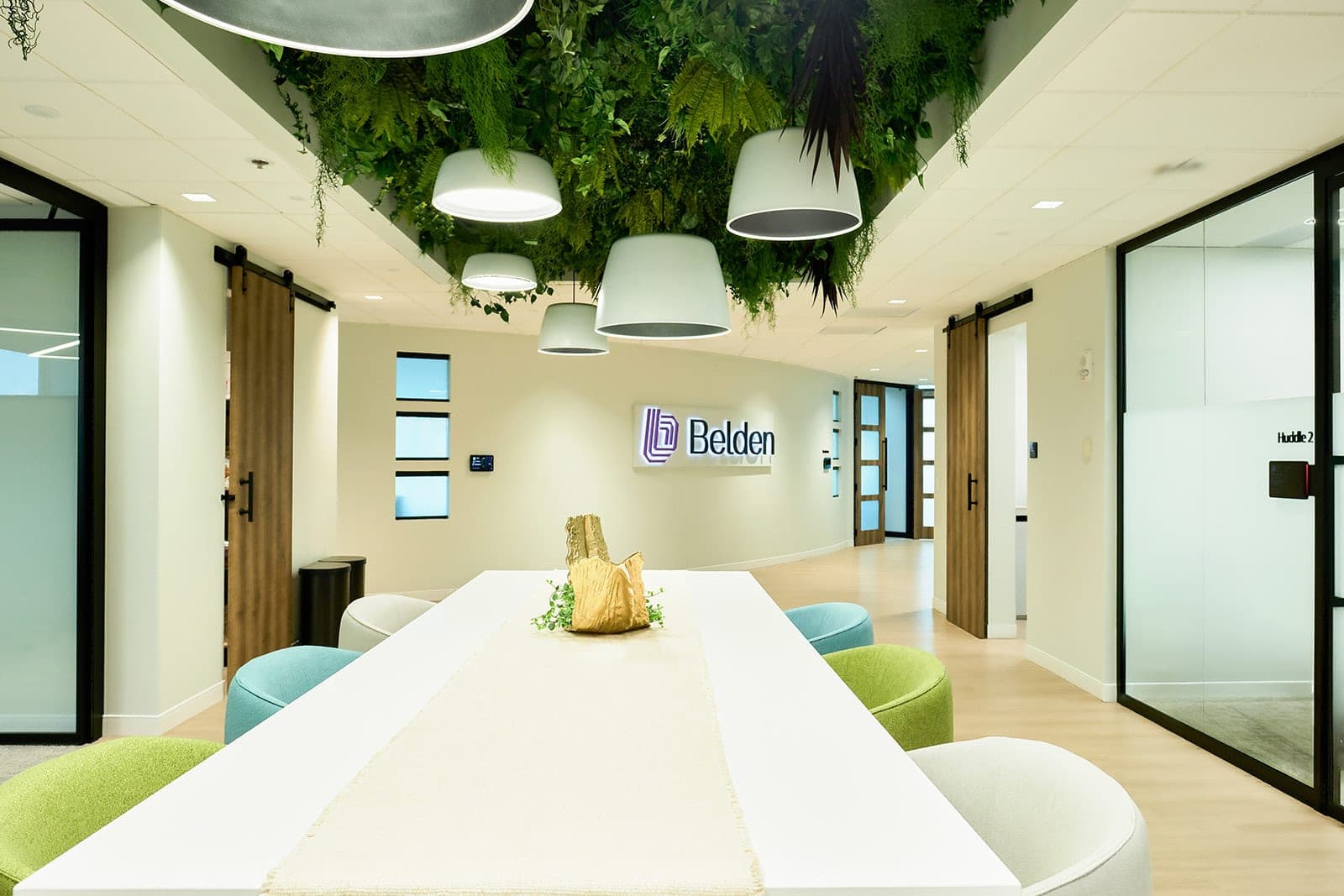 A Belden huddle room in St. Louis with a planted ceiling installation, pendant lamps, a backlit brand sign and a round table with teal and cream chairs, corporate interior photography