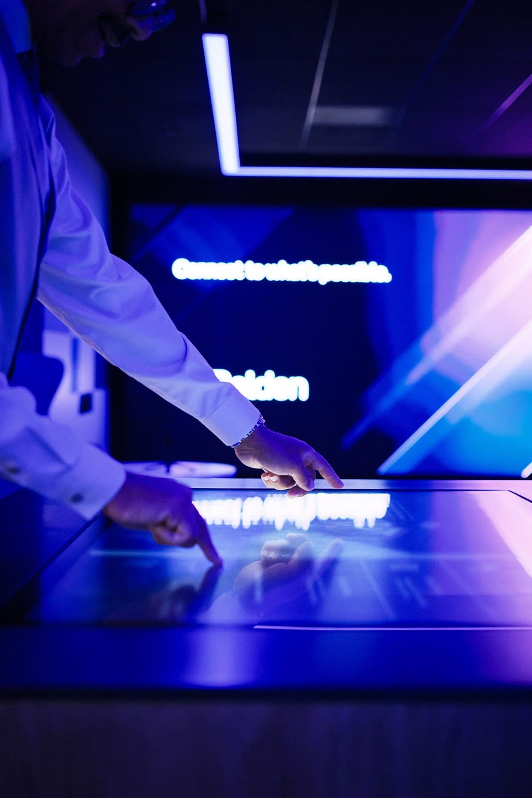 A hand on the Belden interactive touch table with the wall tagline and wordmark legible behind it, detail photography of a technology installation in St. Louis