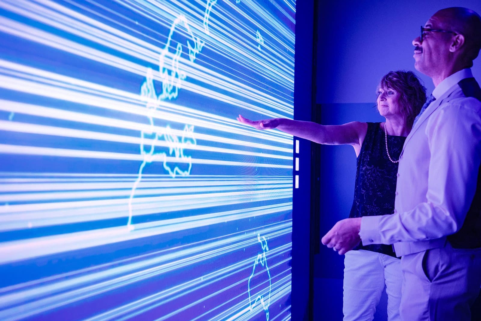 Two colleagues pointing at a global map streaming across the Belden LED video wall, workplace photography in a St. Louis experience center
