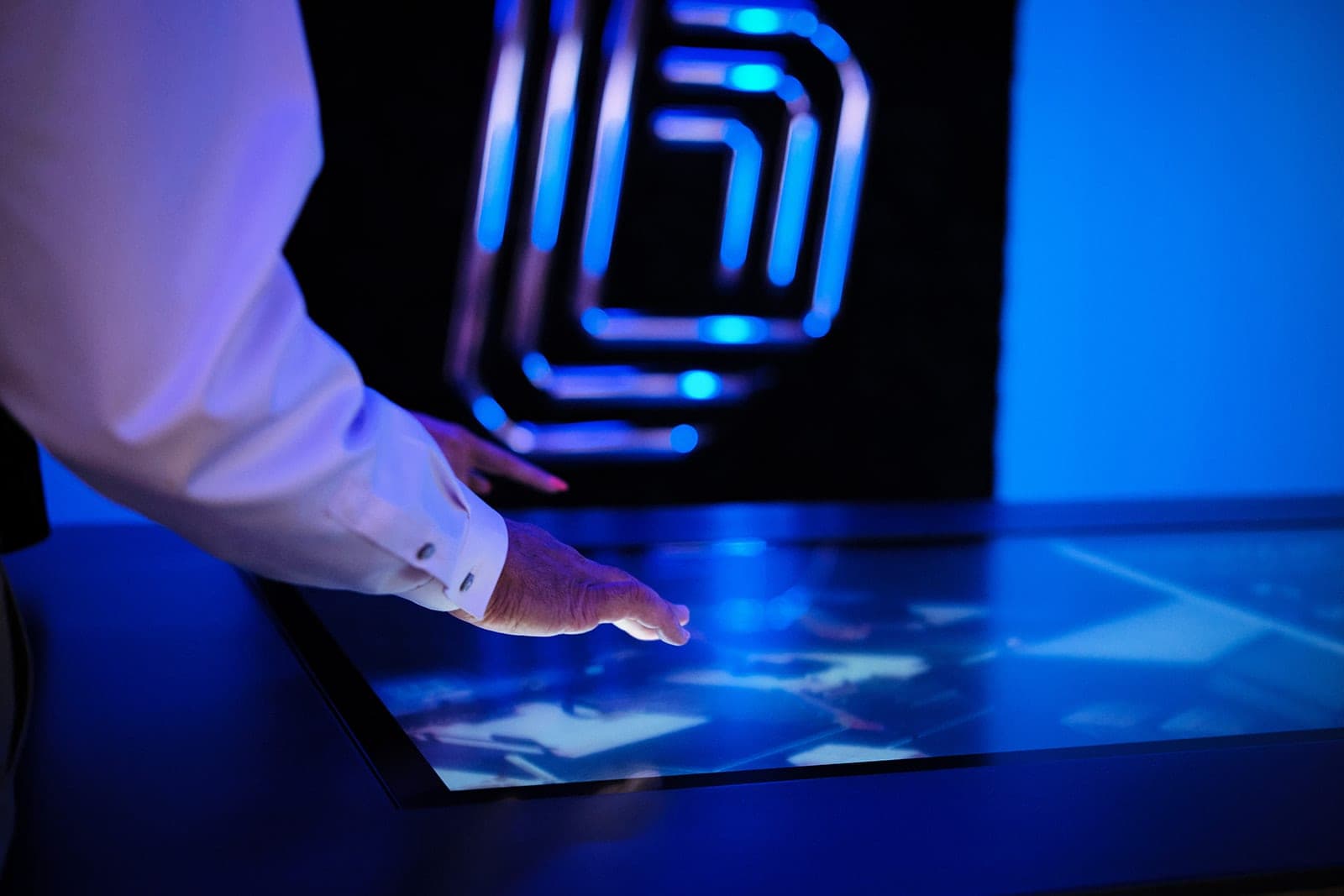 A hand above the glowing touch table screen with the illuminated Belden mark on the moss wall out of focus behind it, macro photography in a dark St. Louis interior