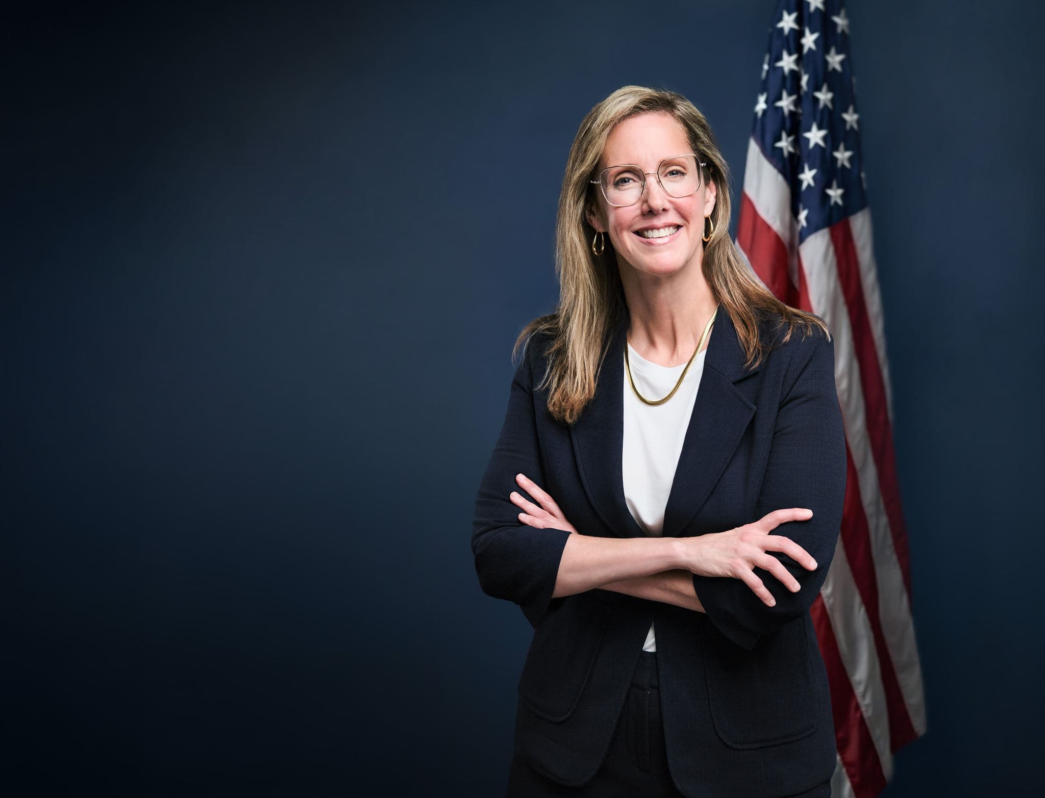 Formal portrait of a public-facing candidate in navy with an American flag background