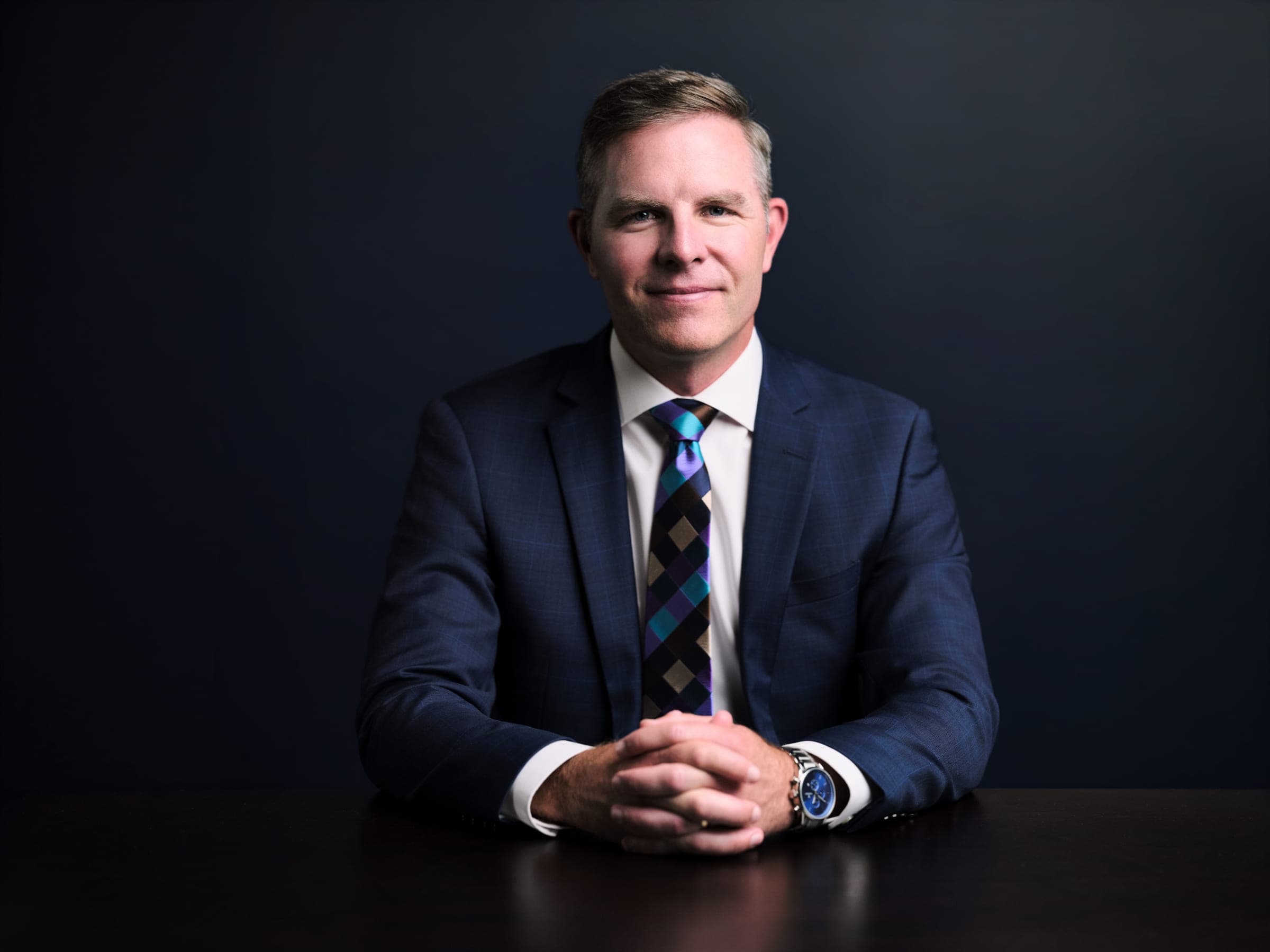 Financial advisor Drew Forrester seated at a dark desk with hands clasped, navy suit and patterned tie, navy studio backdrop, St. Louis personal branding portrait by Henry David