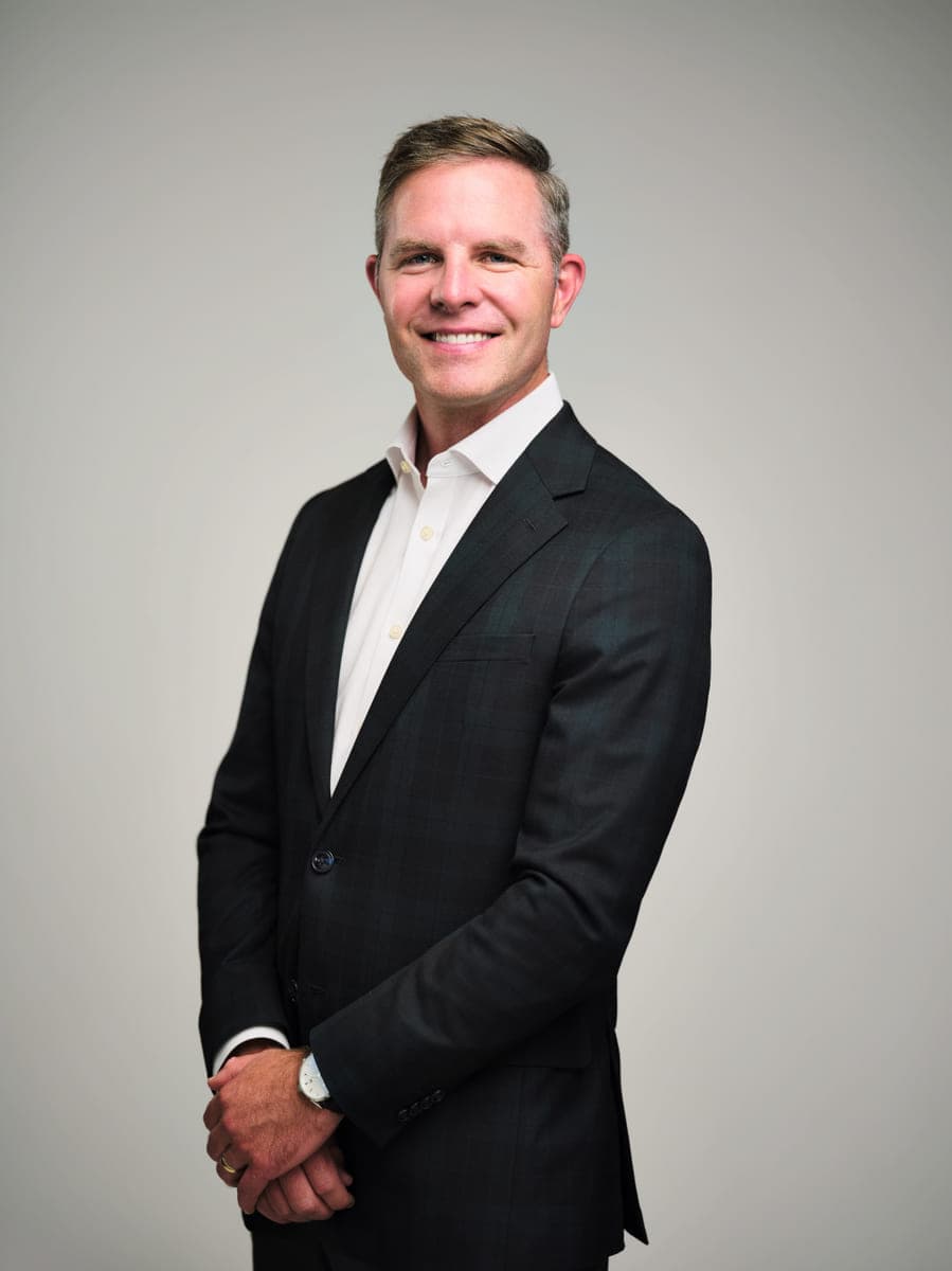 Professional headshot of Drew Forrester in a dark suit and open collar on a grey studio backdrop