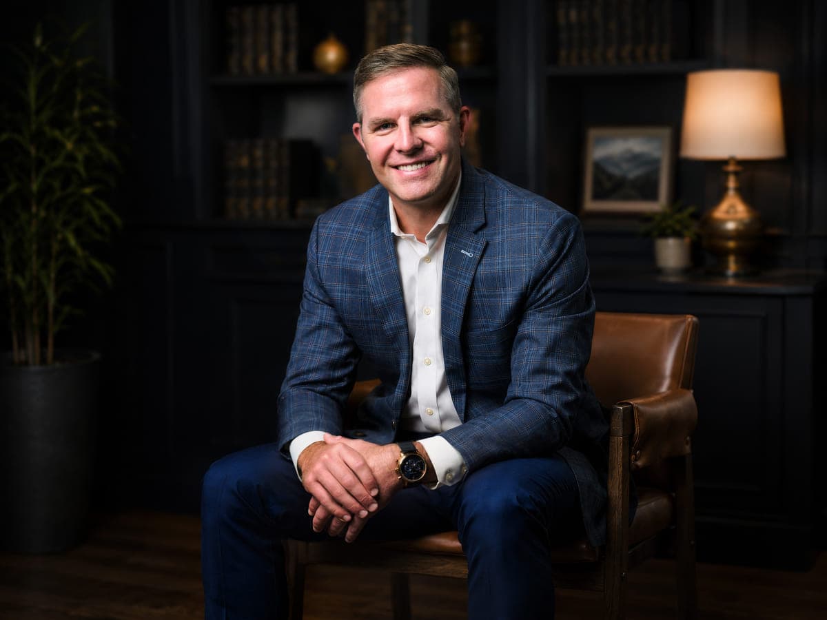 Drew Forrester of Everant Wealth seated in a leather chair in a dark library setting, blue plaid blazer, executive portrait