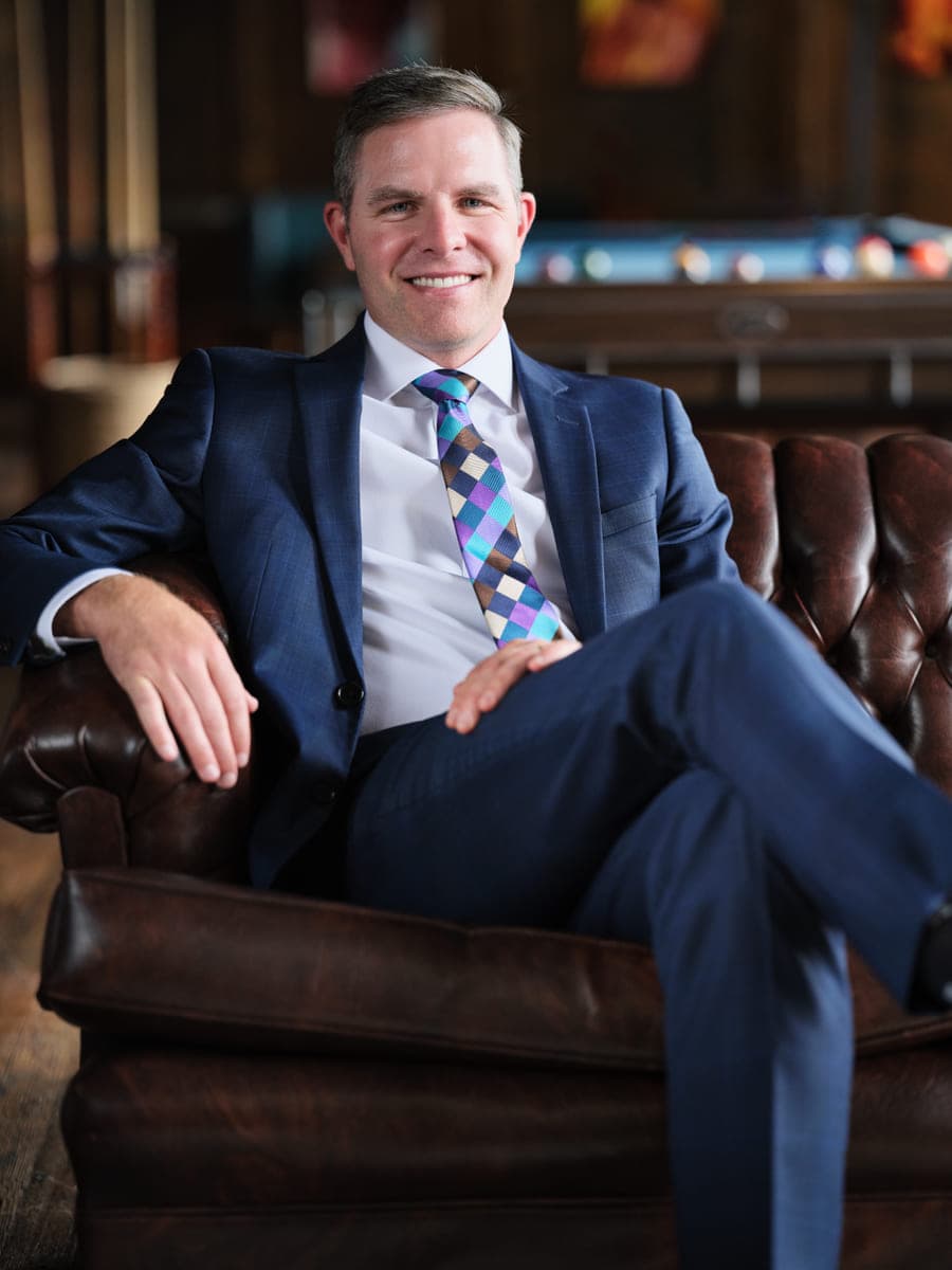 Drew Forrester seated in a tufted leather chair in a navy suit with a pool table behind him, relaxed executive portrait