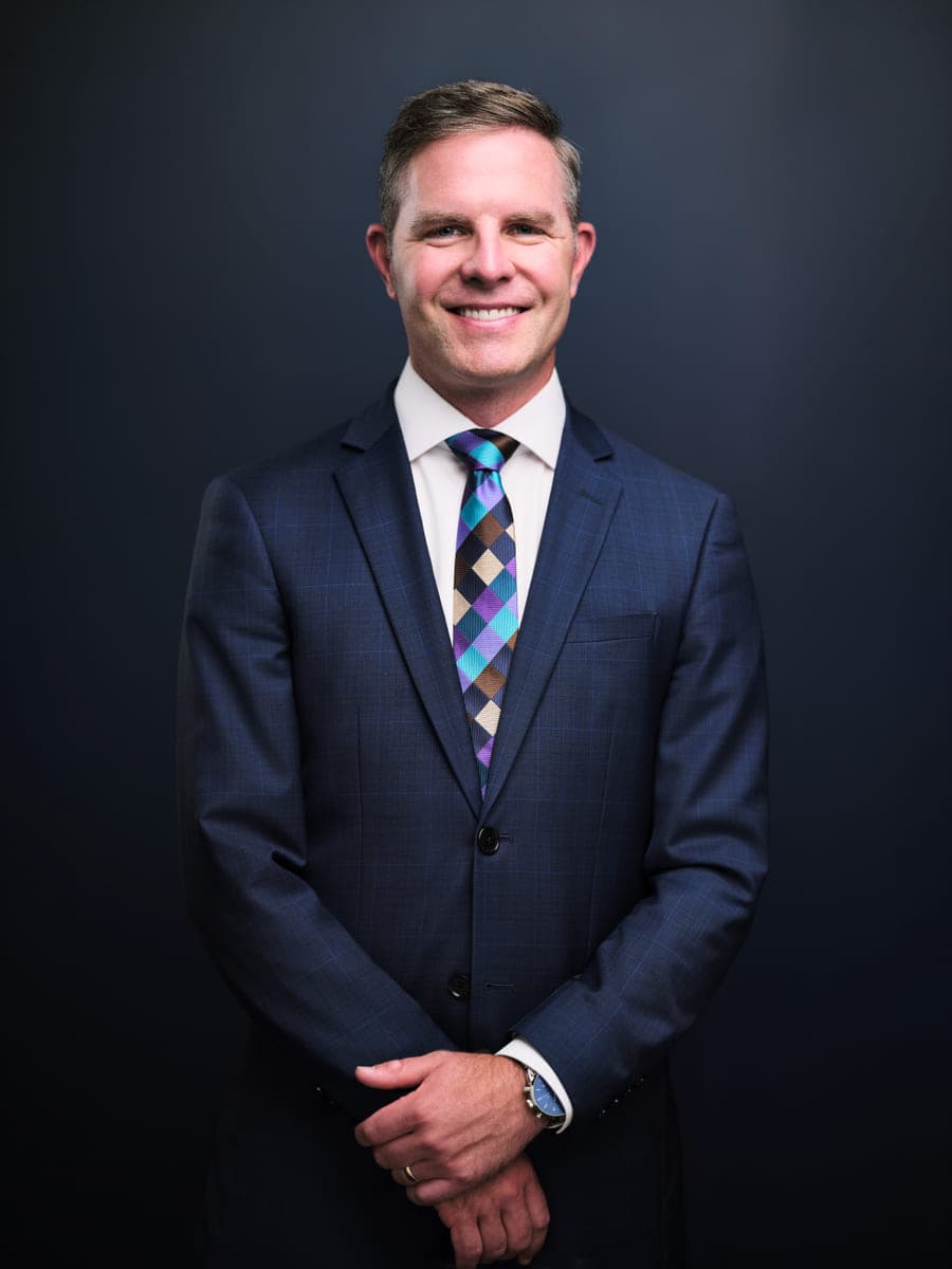 Corporate headshot of Drew Forrester in a navy suit and patterned tie on a navy studio backdrop