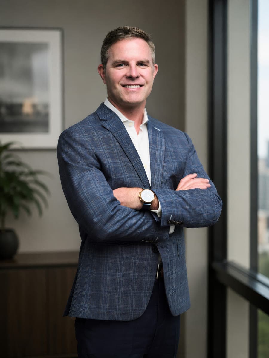 Drew Forrester standing with arms crossed in a blue plaid blazer beside a window in an office setting