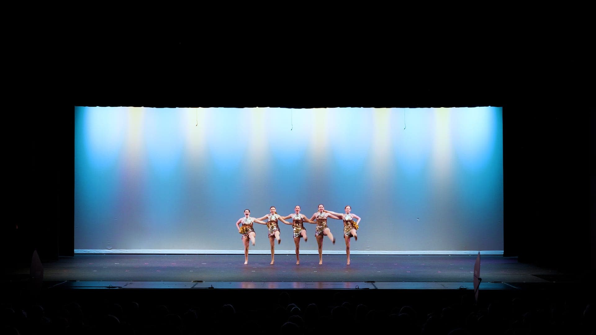 Five dancers in gold sequins linked in a kick line on a St. Louis dance recital stage