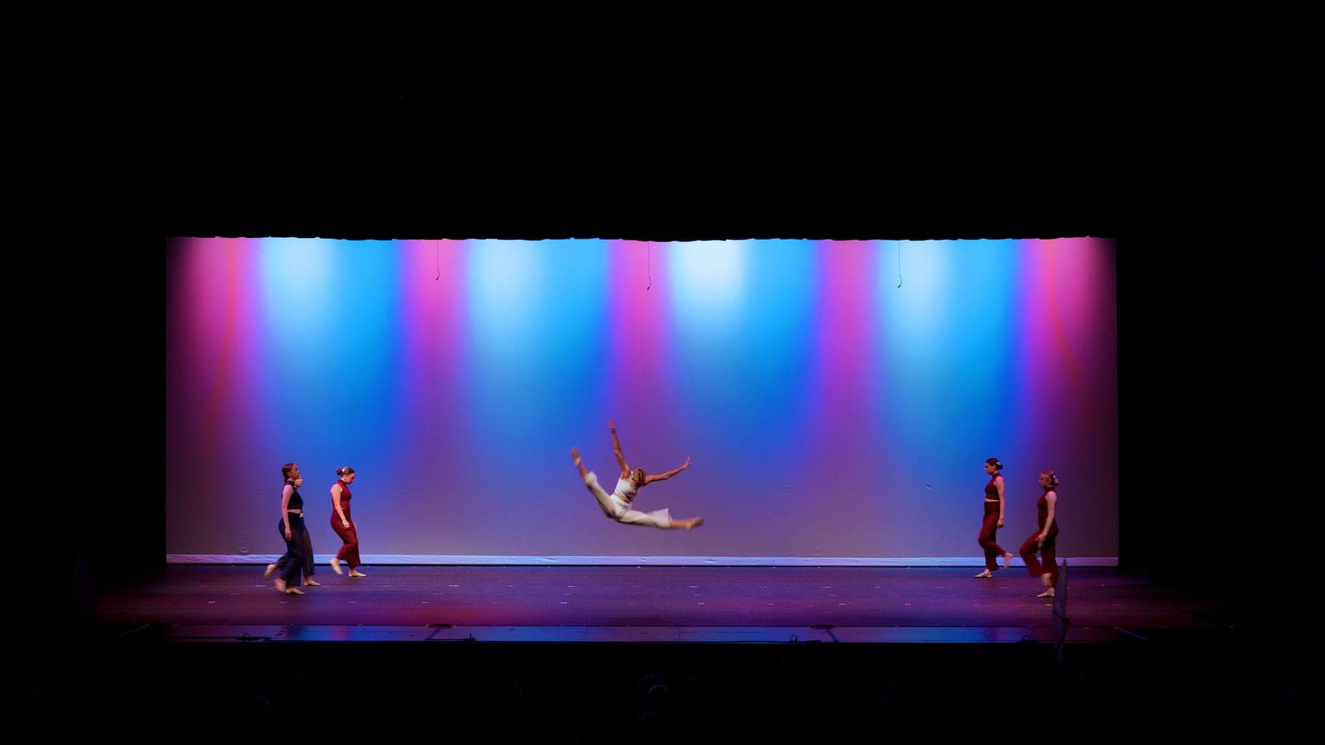 A dancer caught mid-leap with legs extended at a St. Louis dance recital under a blue backdrop