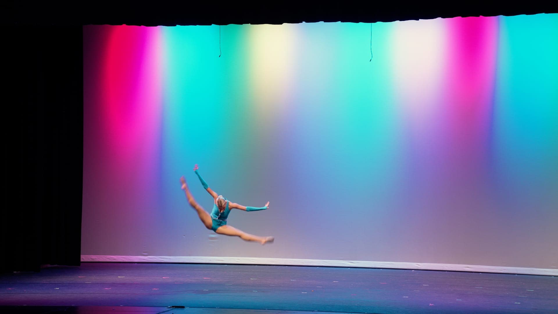 Solo dancer in teal leaps in a full split against a rainbow backdrop at a St. Louis dance recital