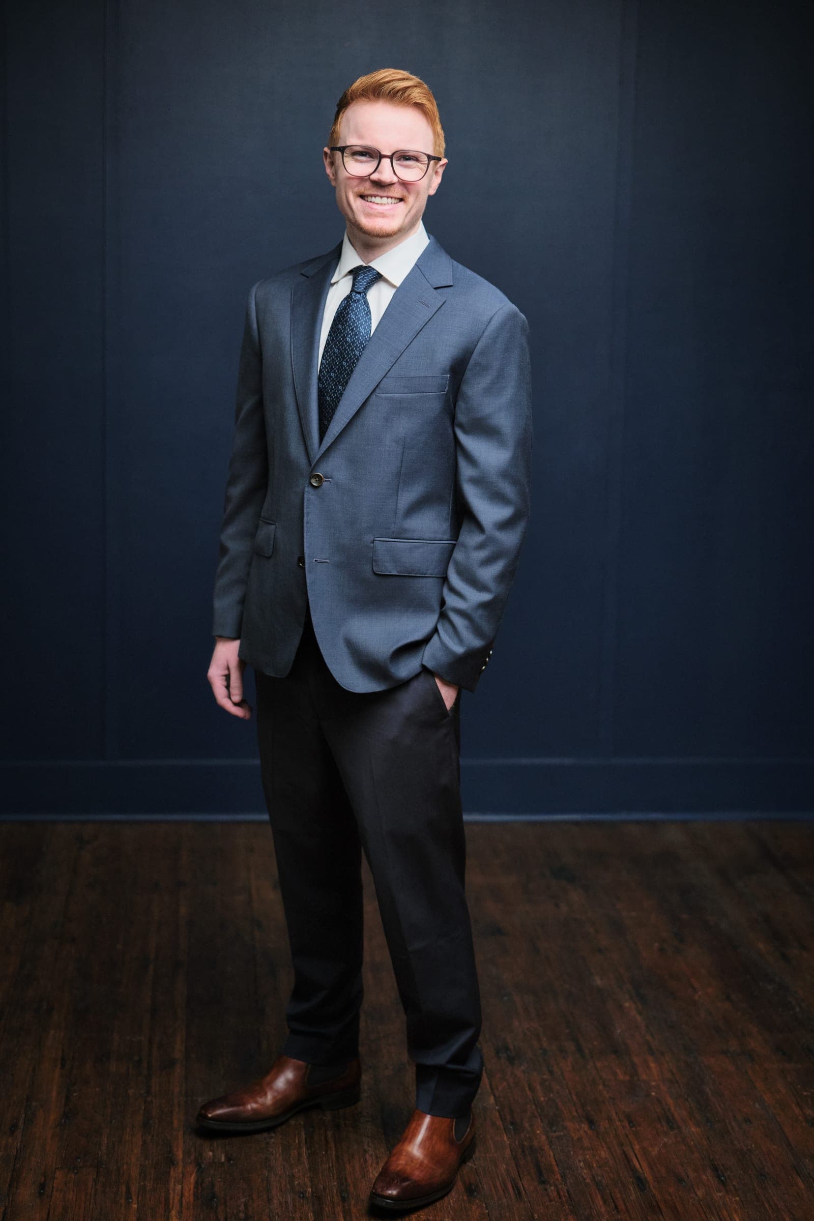 Full-length portrait of an attorney at The Dixon Injury Firm in a blue suit and glasses, hand in pocket, on a navy backdrop over a wood floor in St. Louis by Henry David Photography