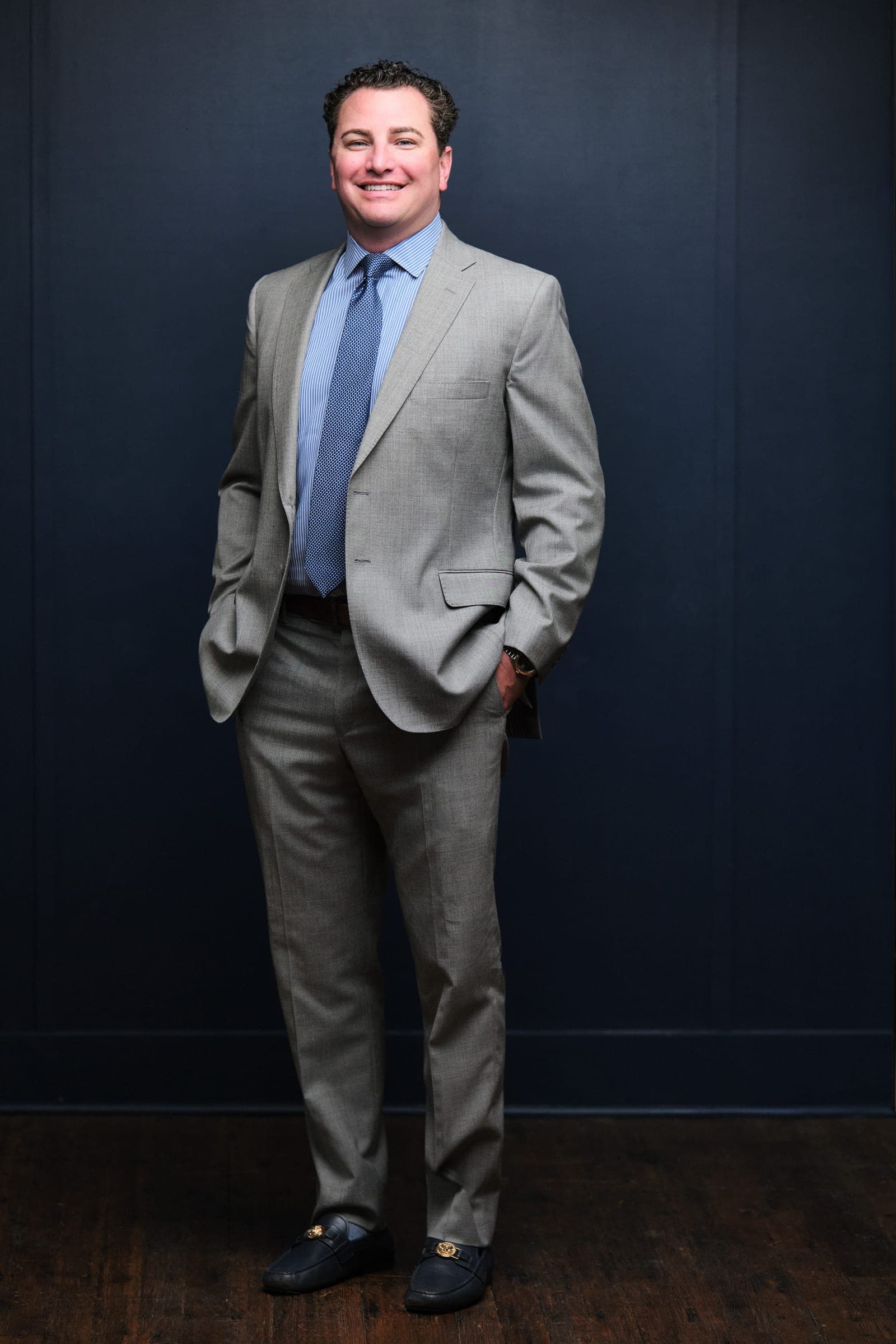 Full-length portrait of an attorney at The Dixon Injury Firm in a light gray suit, hands in pockets, on a navy backdrop over a wood floor in St. Louis by Henry David Photography