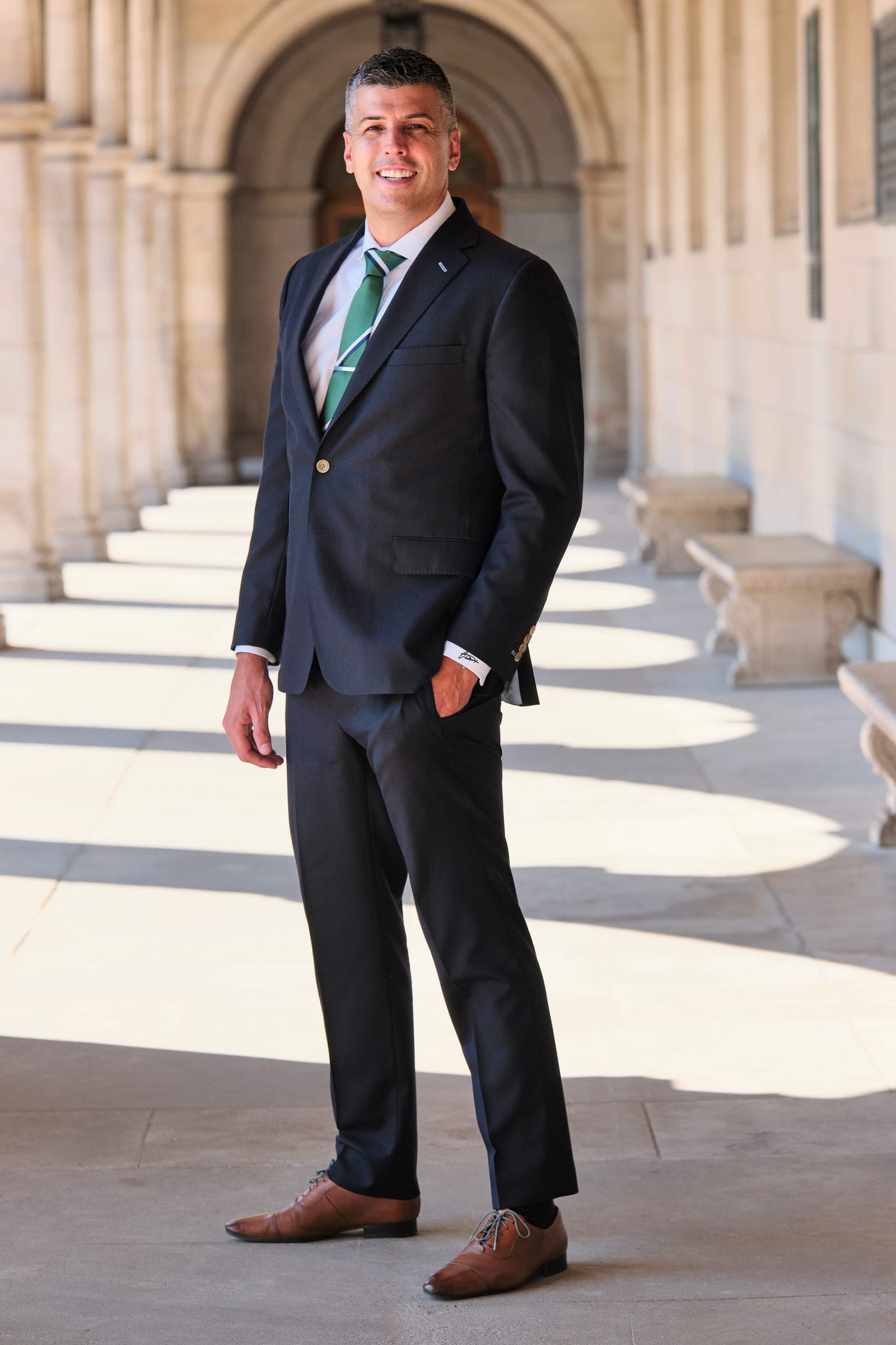 Attorney at The Dixon Injury Firm in a dark suit standing in the sunlit stone arcade of Brookings Hall at Washington University in St. Louis, portrait by Henry David Photography