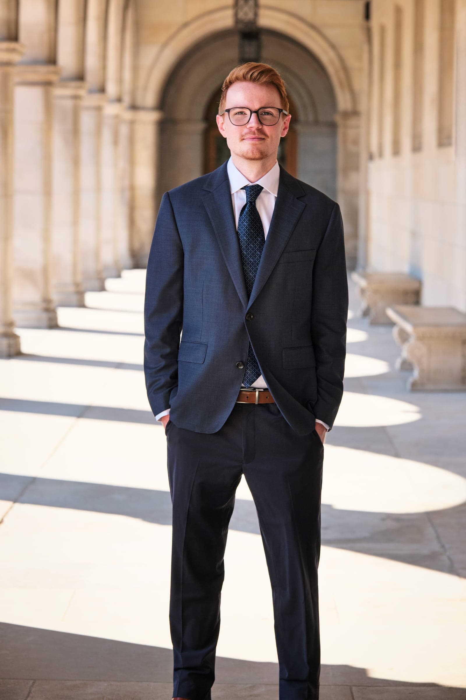 Attorney at The Dixon Injury Firm in a navy suit and glasses, hands in pockets, in the stone arcade of Brookings Hall at Washington University in St. Louis by Henry David Photography