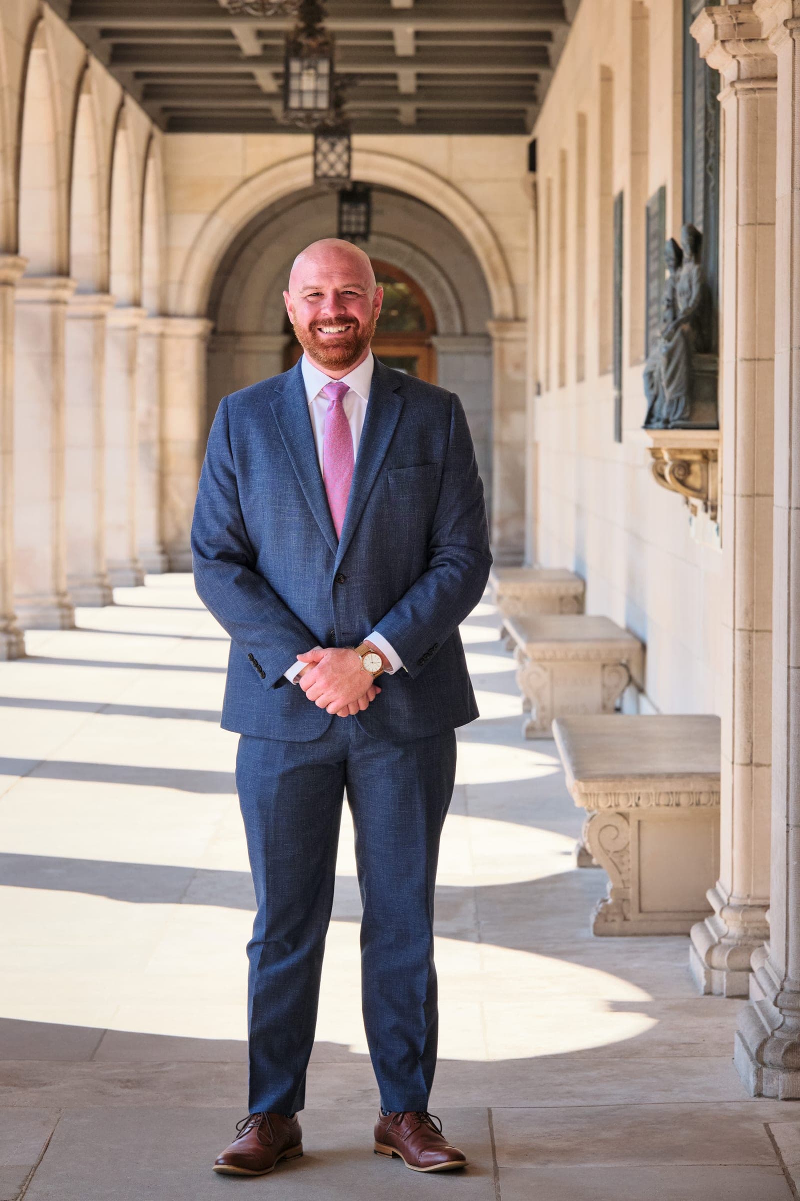 Attorney at The Dixon Injury Firm in a blue suit and pink tie, hands clasped, in the stone arcade of Brookings Hall at Washington University in St. Louis by Henry David Photography