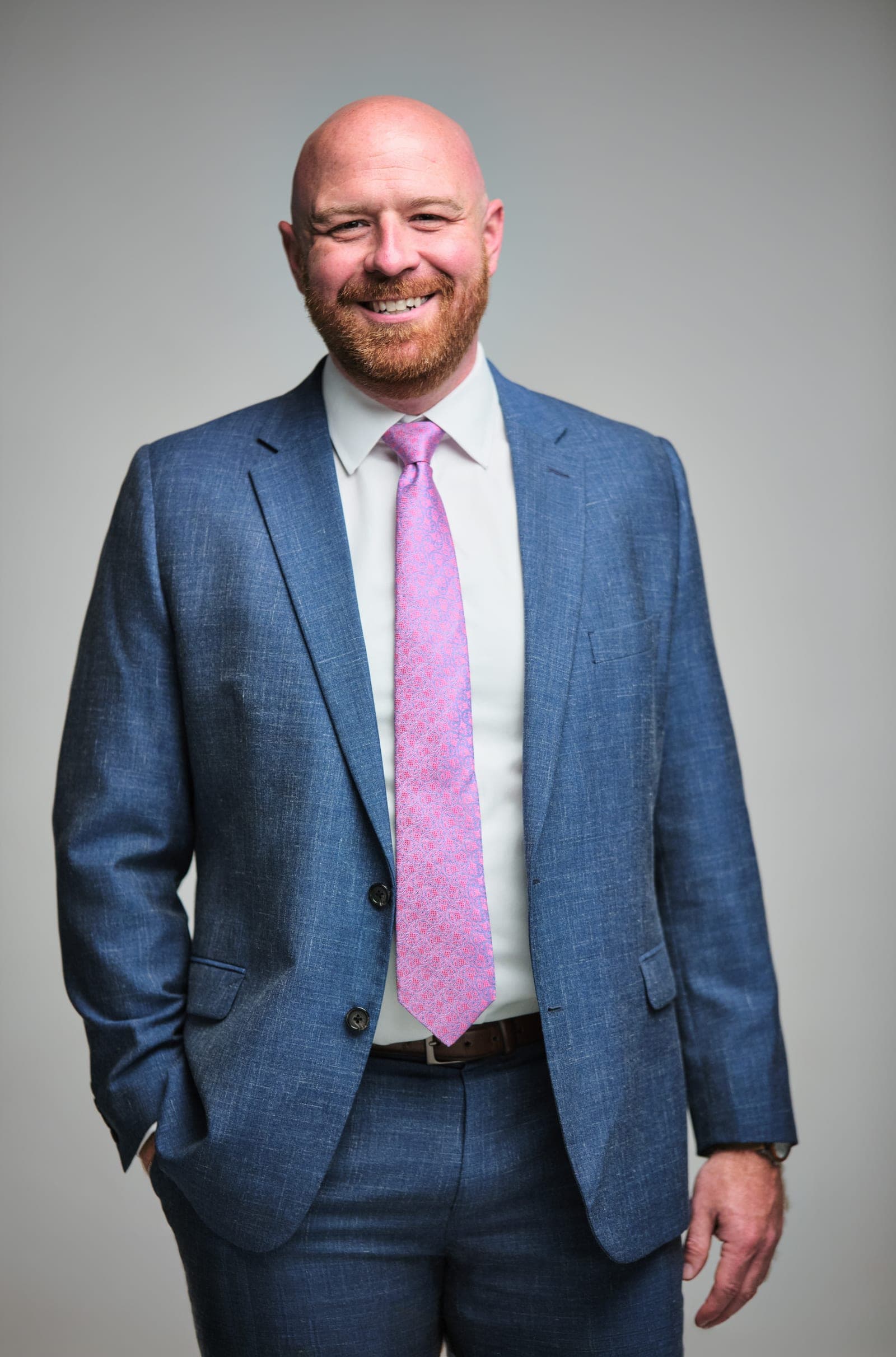 Attorney at The Dixon Injury Firm smiling with one hand in a pocket, light blue suit and pink tie on a light gray backdrop, St. Louis portrait by Henry David Photography