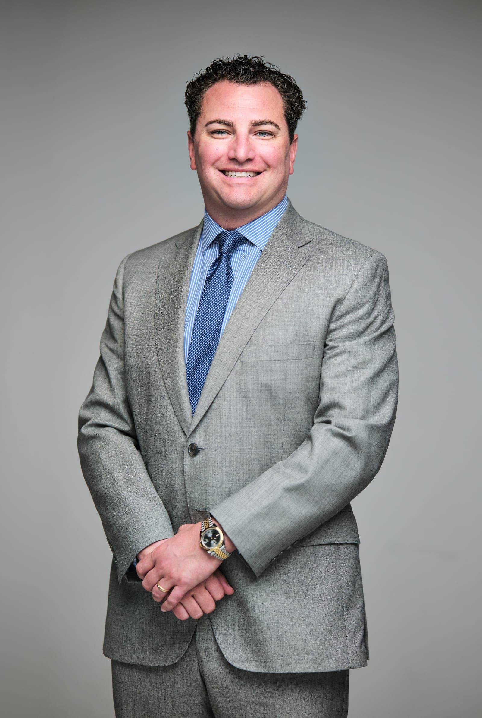 Attorney at The Dixon Injury Firm in a light gray suit and blue tie, hands clasped, smiling, portrait on a light gray backdrop in St. Louis by Henry David Photography