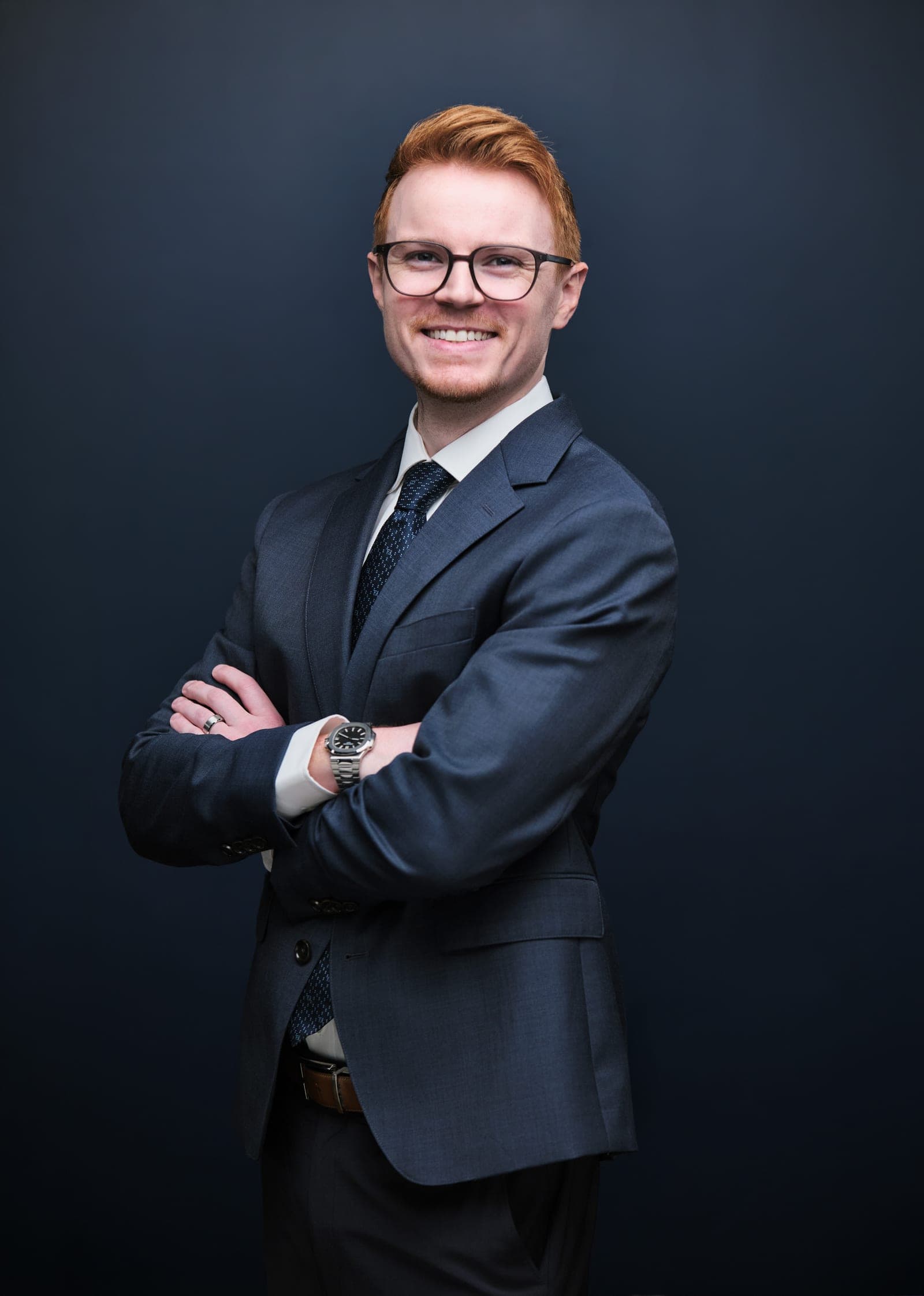 Attorney at The Dixon Injury Firm in glasses and a navy suit, body angled with arms crossed, smiling, navy backdrop portrait in St. Louis by Henry David Photography