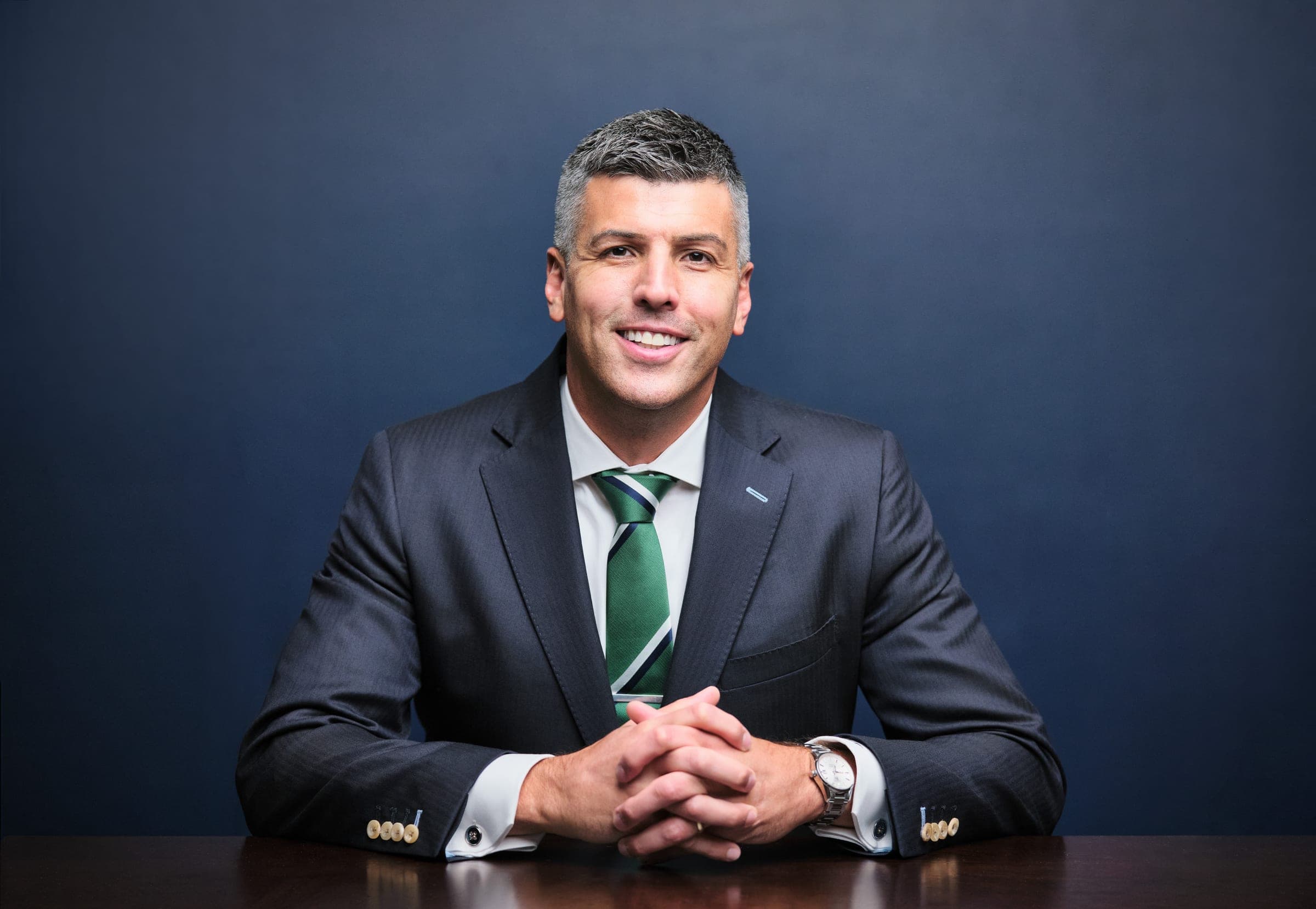 Attorney at The Dixon Injury Firm seated at a dark wood table with hands folded, dark suit and green tie, navy backdrop, St. Louis law firm portrait by Henry David Photography
