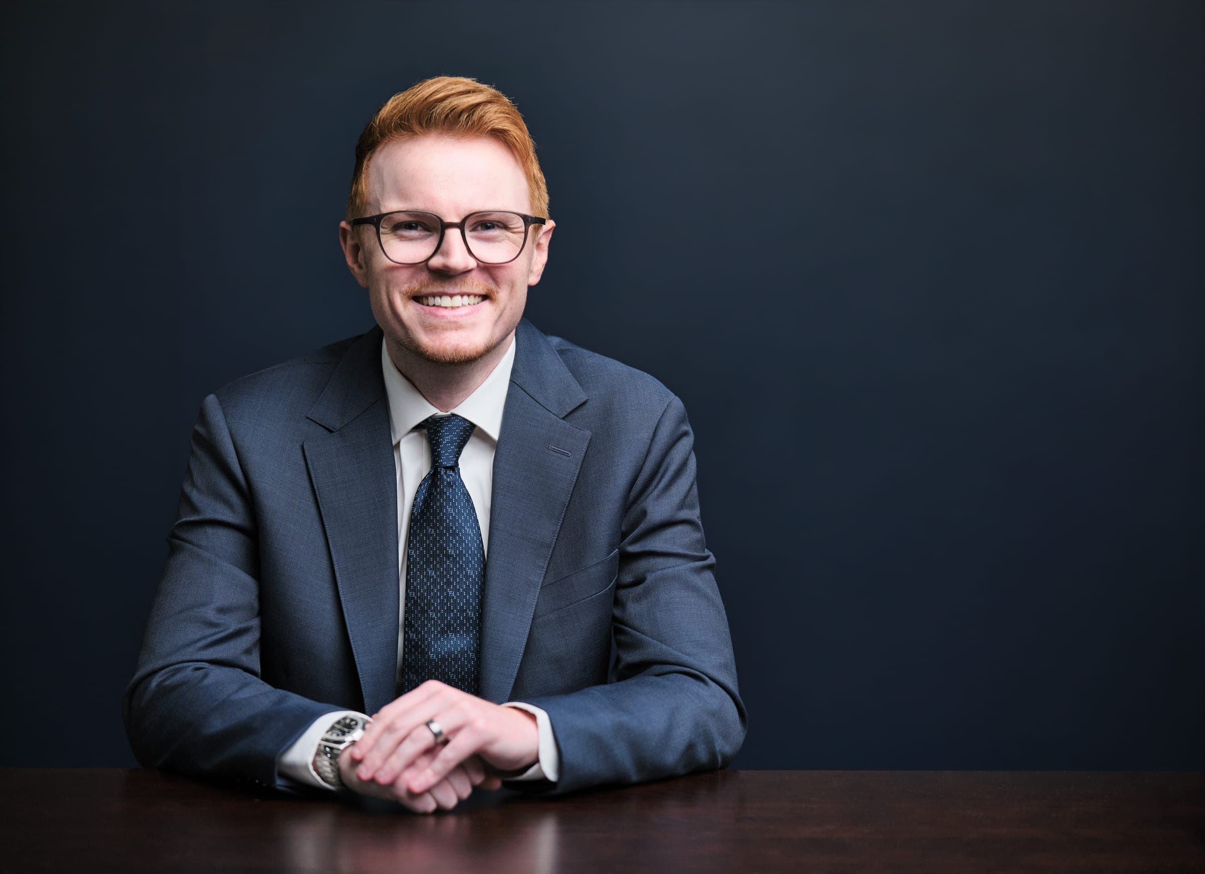 Attorney at The Dixon Injury Firm in a navy suit and glasses, seated at a dark wood table with hands folded, navy backdrop, St. Louis law firm portrait by Henry David Photography