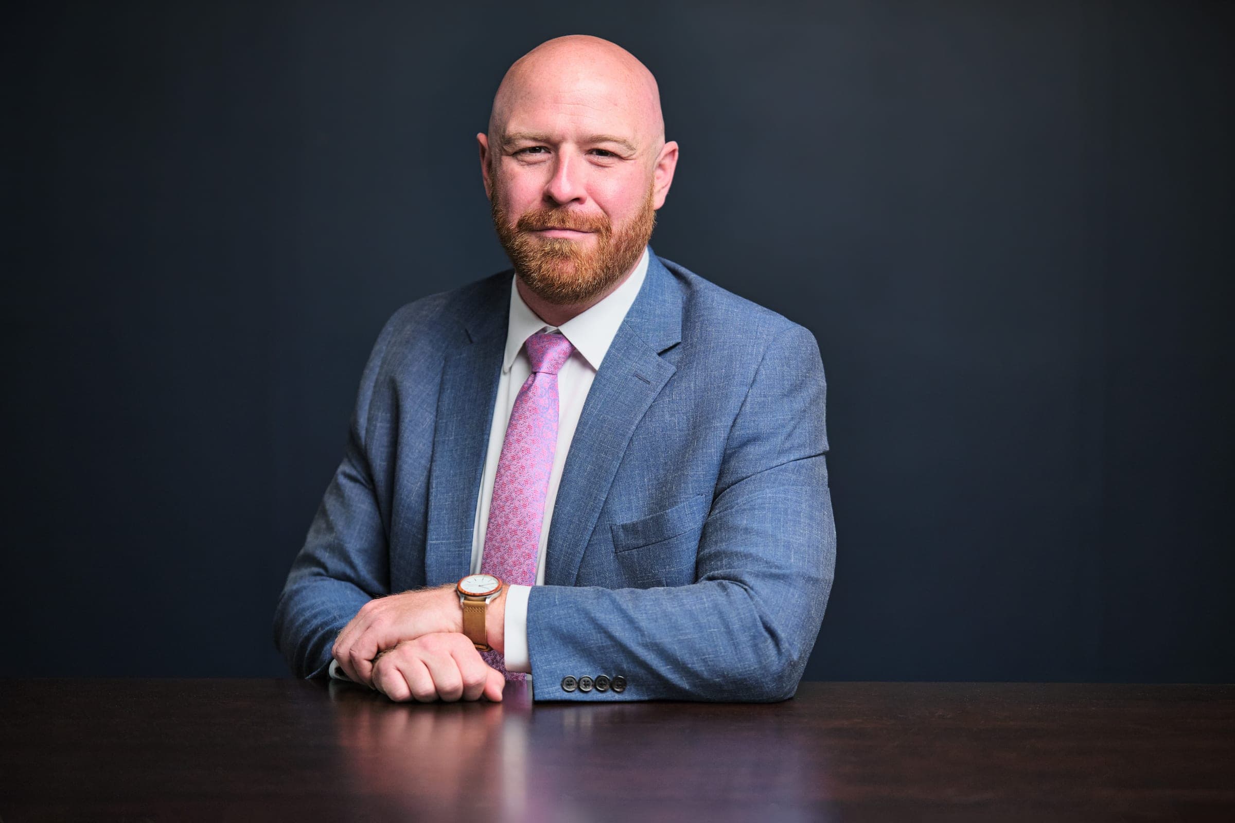 Attorney at The Dixon Injury Firm in a light blue suit and pink tie, seated at a dark wood table, navy backdrop, St. Louis law firm portrait by Henry David Photography