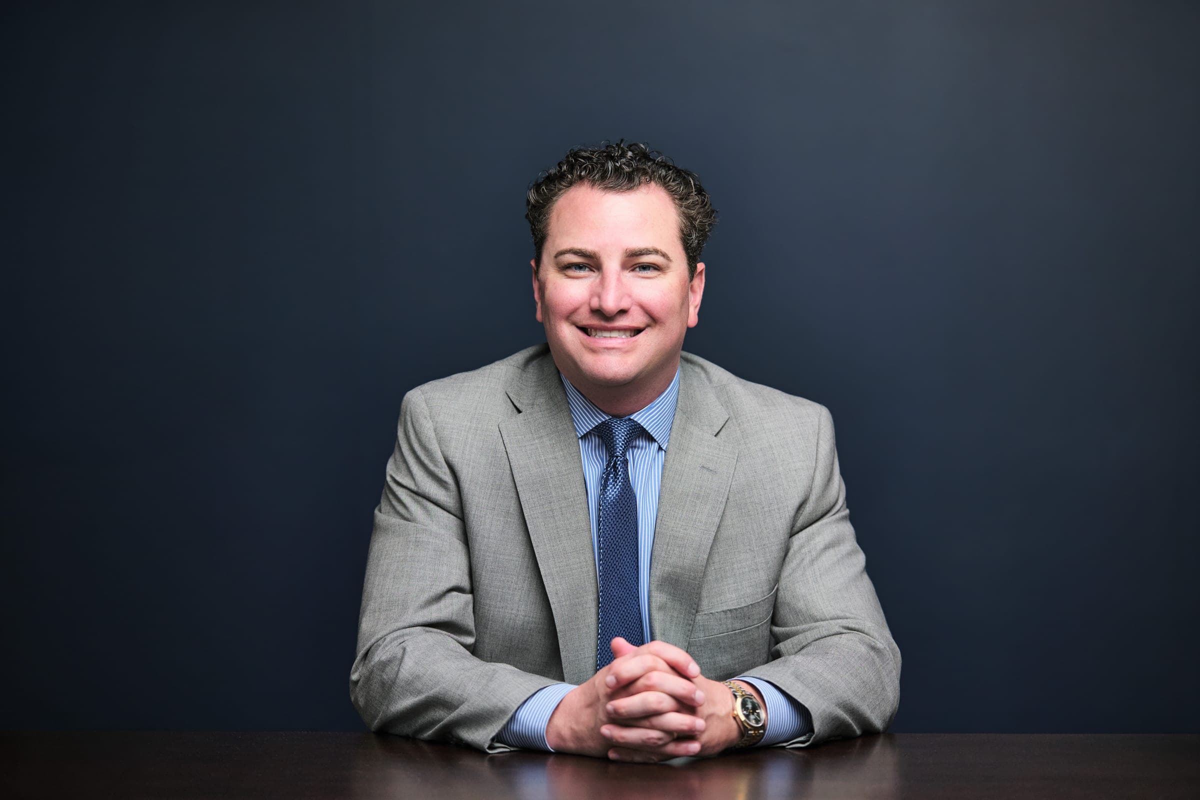 Attorney at The Dixon Injury Firm in a light gray suit and navy tie, seated at a dark wood table with hands folded, navy backdrop, St. Louis by Henry David Photography