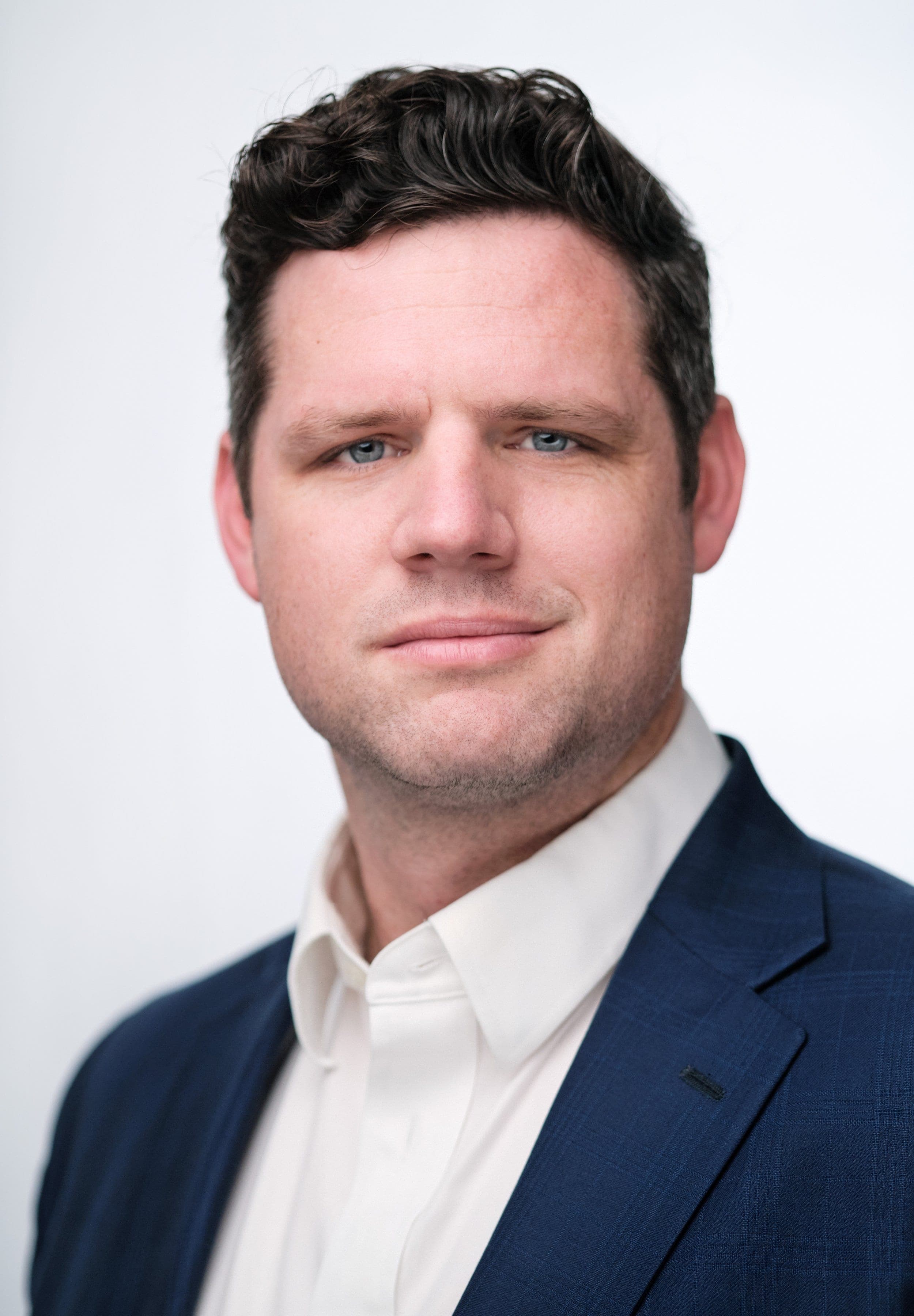Man in a navy suit and open-collar white shirt on a light backdrop, photographed at our headshot booth at a business expo