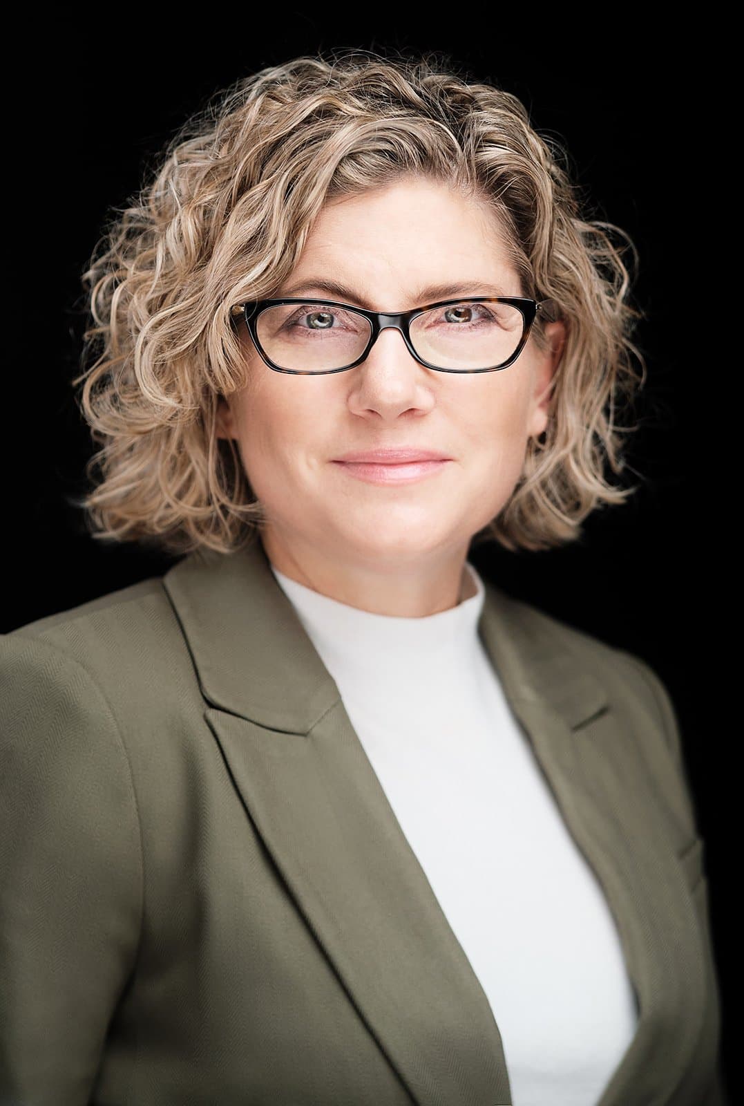 Woman with curly hair and glasses in an olive blazer, photographed at our headshot booth at a conference