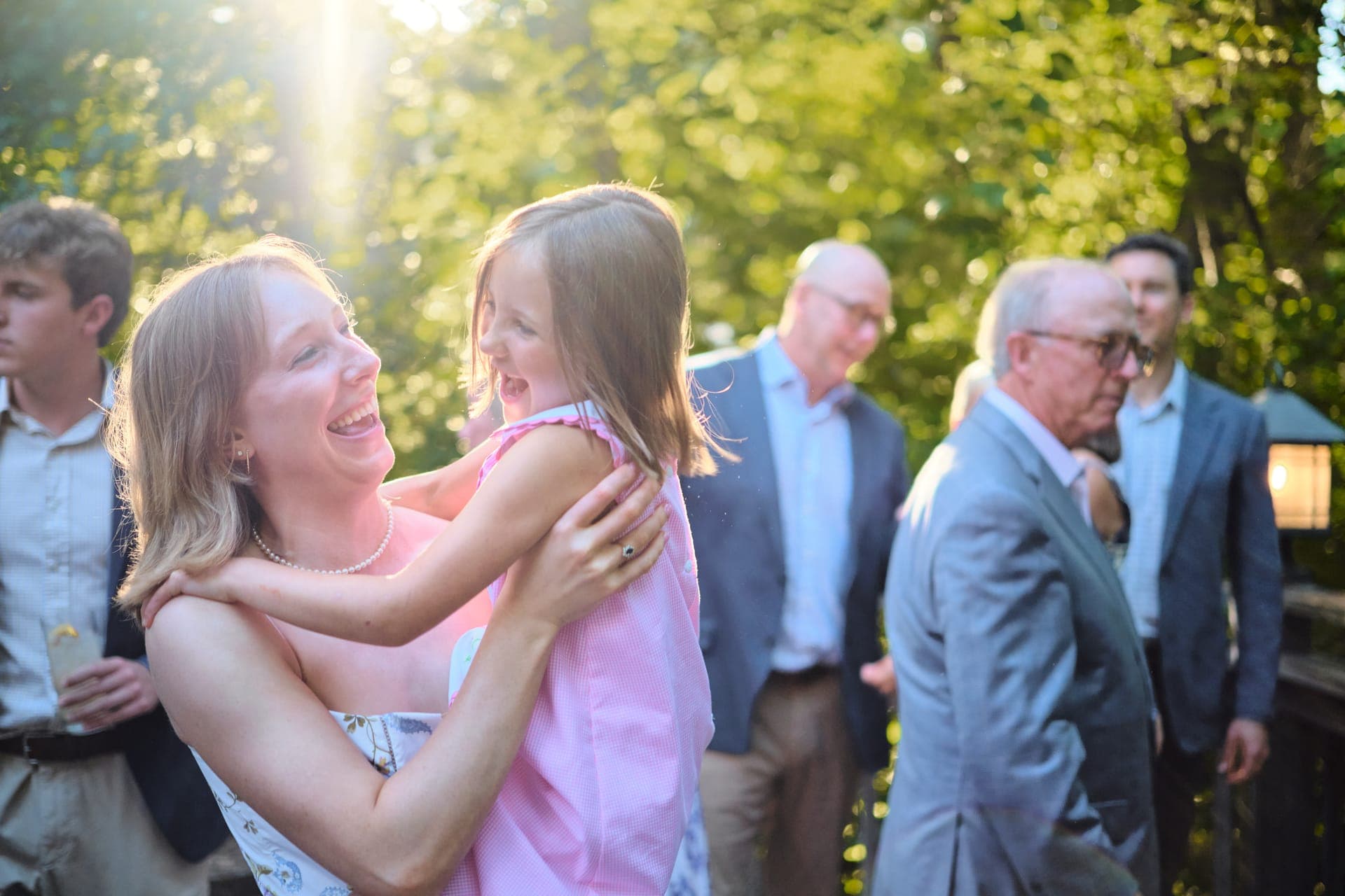 A mother lifts her laughing daughter into a hug in golden evening light at a family reunion