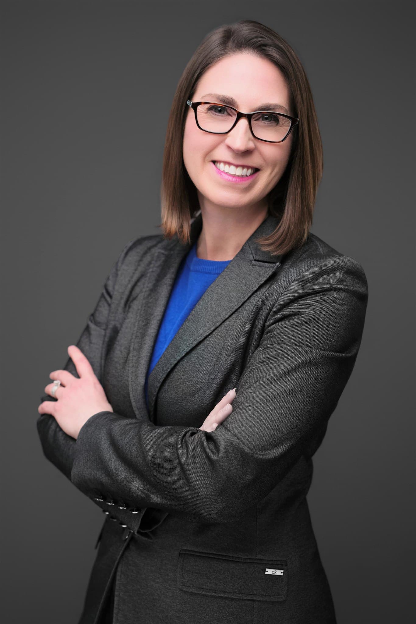 Approachable professional woman in gray blazer and blue top with glasses, arms crossed, against a dark gray backdrop by Henry David Photography