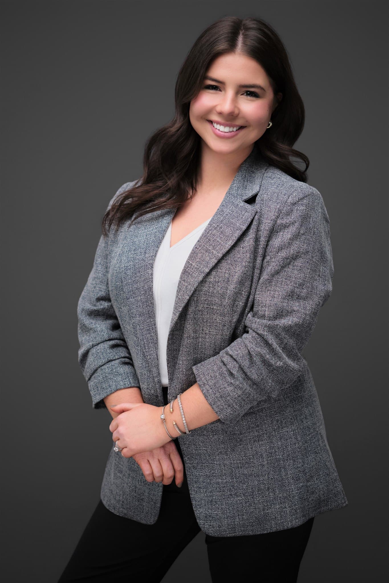 Warm, approachable woman in gray tweed blazer and white top with dark wavy hair against a dark gray backdrop by Henry David Photography
