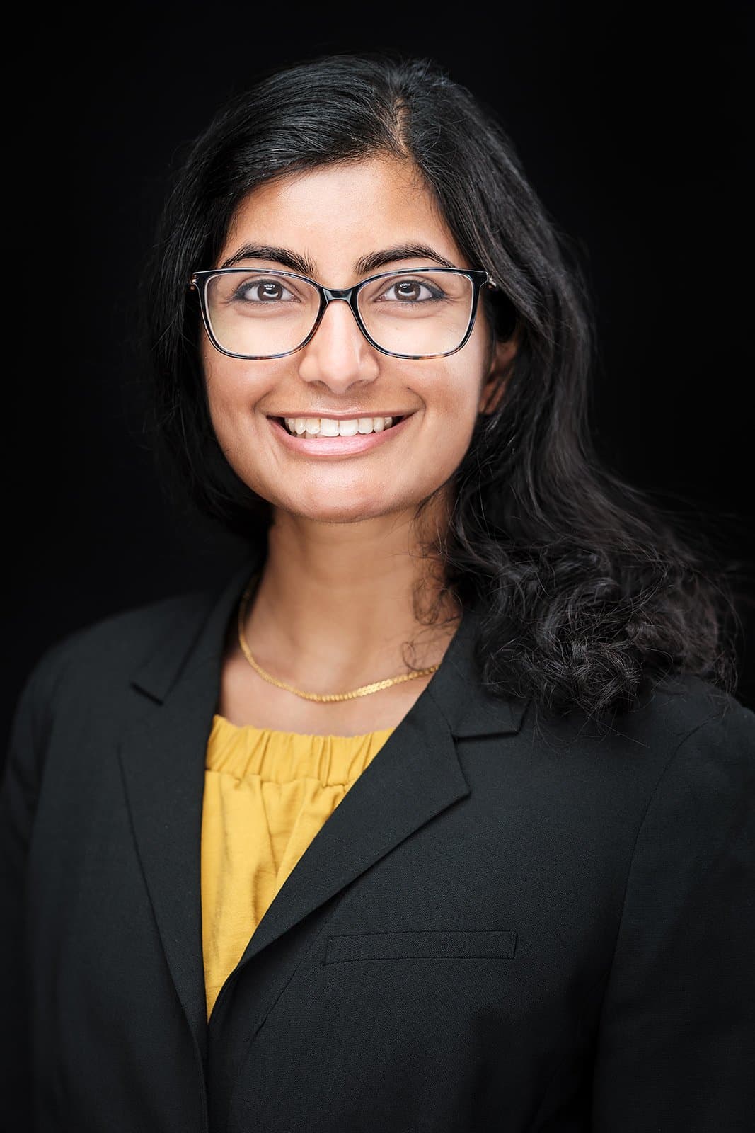 Smiling woman in glasses, black blazer, yellow top, black backdrop by Henry David Photography, the standard every Central West End headshot session is held to