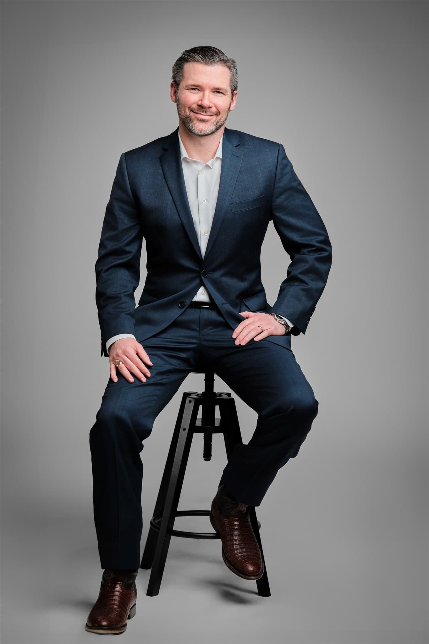 Confident executive man in dark navy suit seated on stool with relaxed smile against a light gray backdrop by Henry David Photography