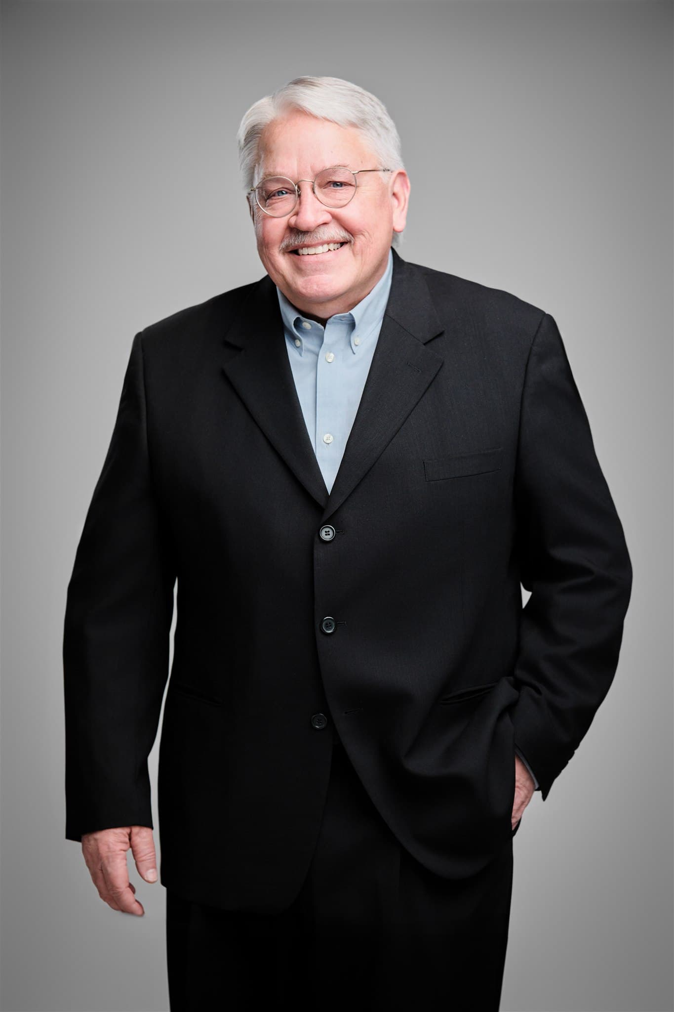 Friendly senior professional man in black suit and light blue shirt with glasses against a light gray backdrop by Henry David Photography