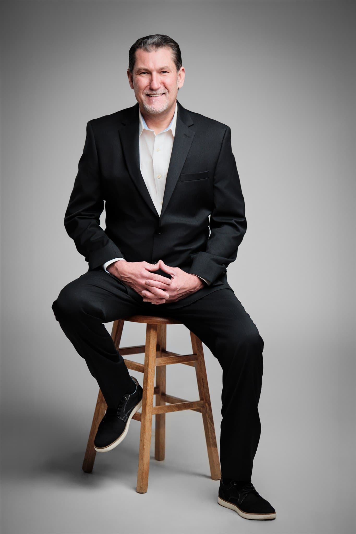 Confident professional man in black suit and open-collar white shirt seated on wooden stool against a light gray backdrop by Henry David Photography