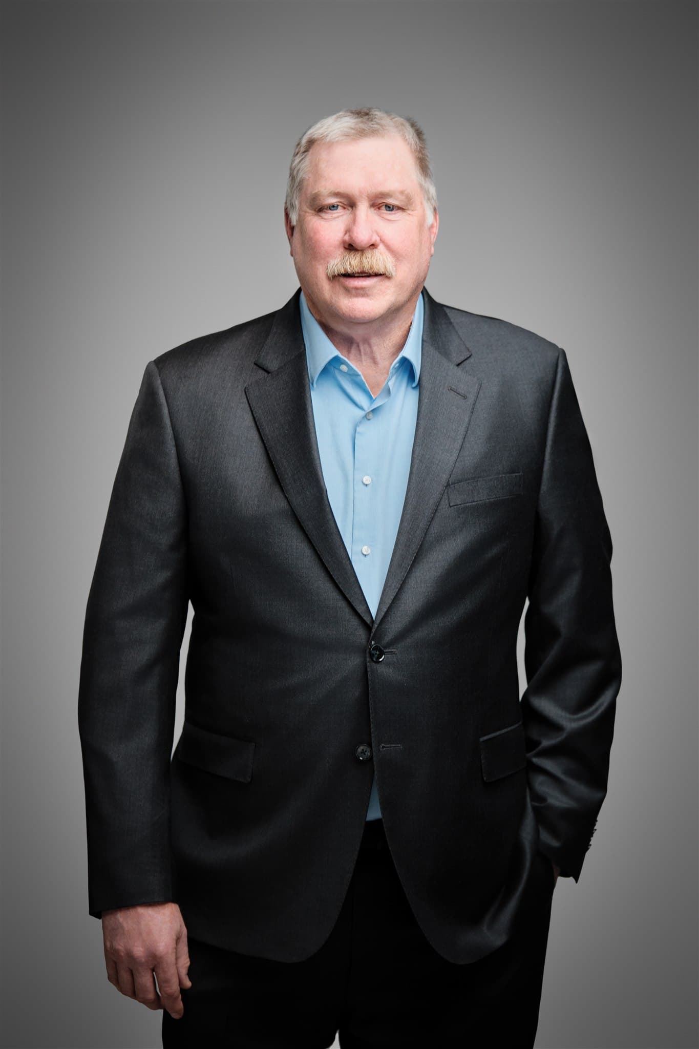 Composed senior executive man in charcoal blazer and light blue shirt with mustache against a light gray backdrop by Henry David Photography