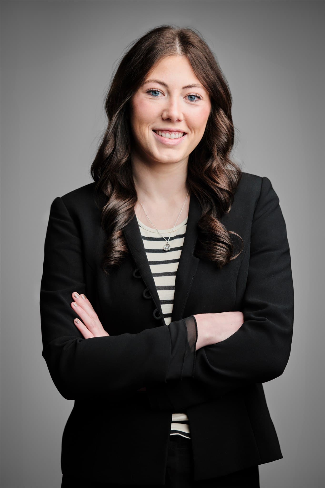 Confident young woman in black blazer and striped top with arms crossed against a light gray backdrop by Henry David Photography