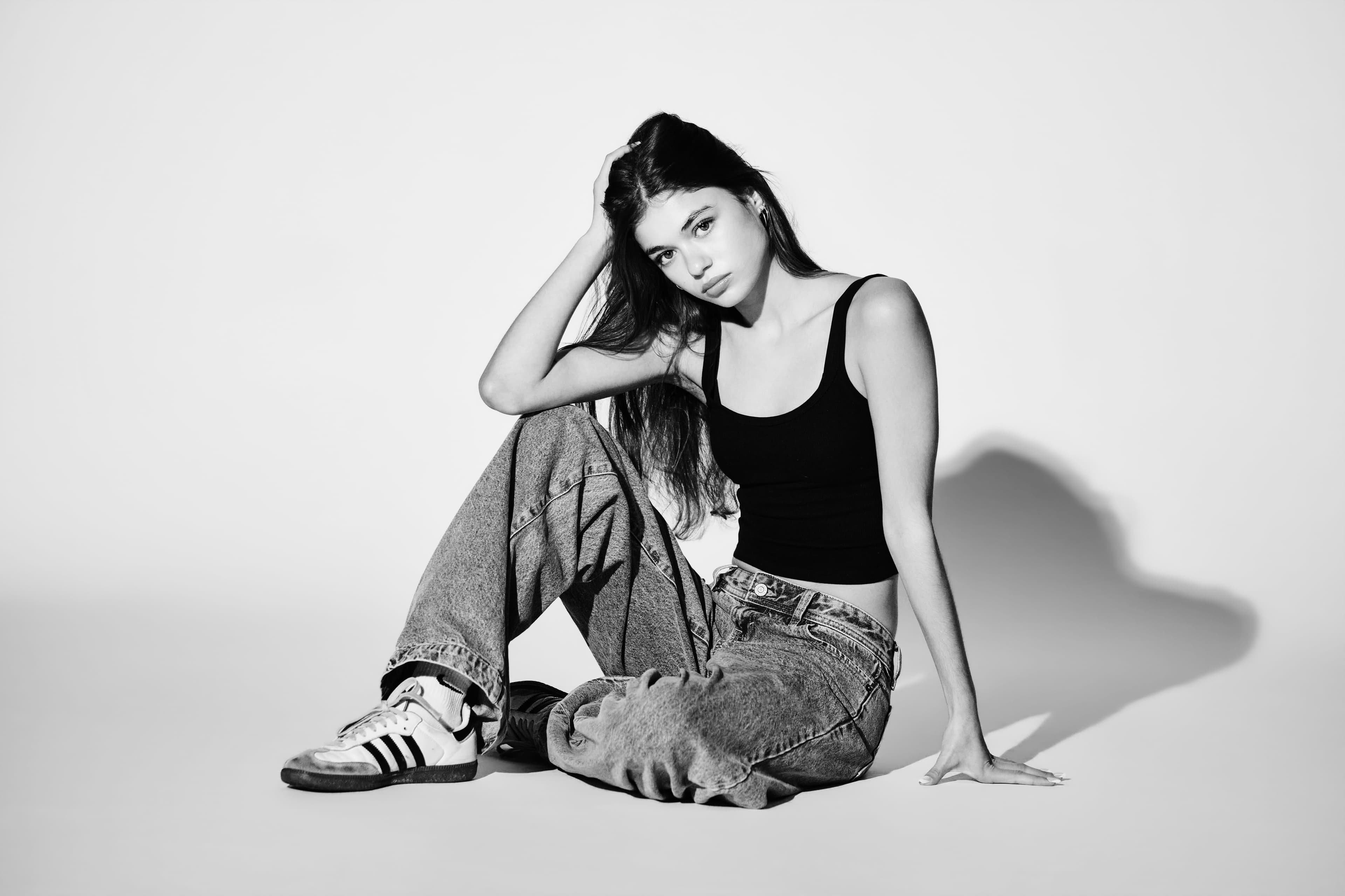 Emme Starkey seated on the studio floor with a hand in her hair, black tank top and jeans, hard light, black and white