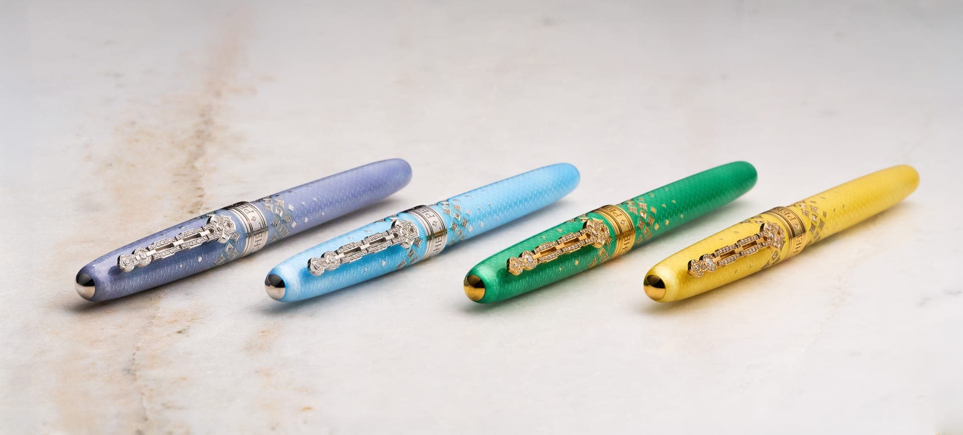 The diamond-set caps of the four Michel Perchin Majesty colorways lined up in a row on marble, each barrel finished in guilloche enamel